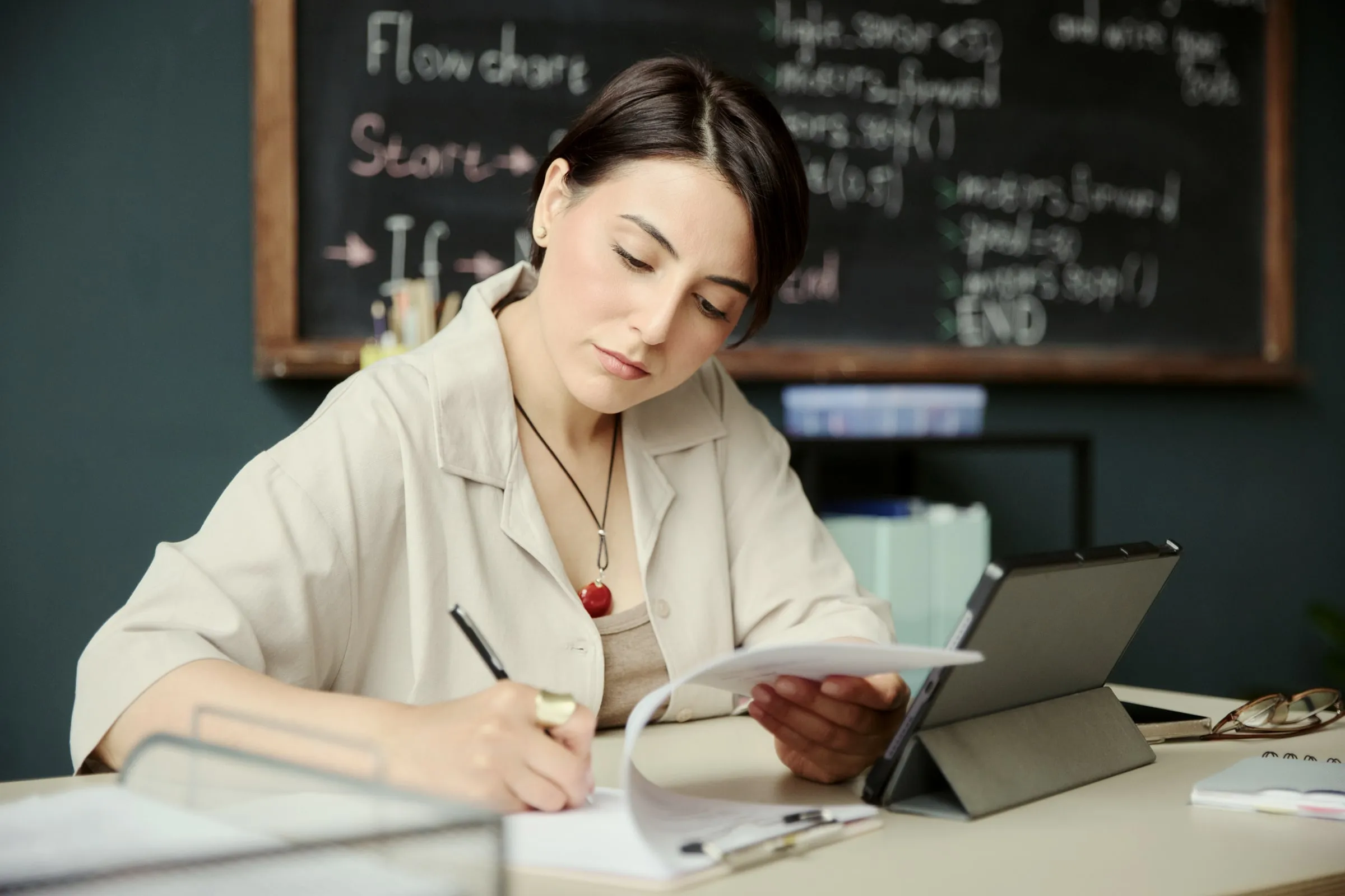 Woman at a desk writing notes beside a tablet in a small office with a chalkboard