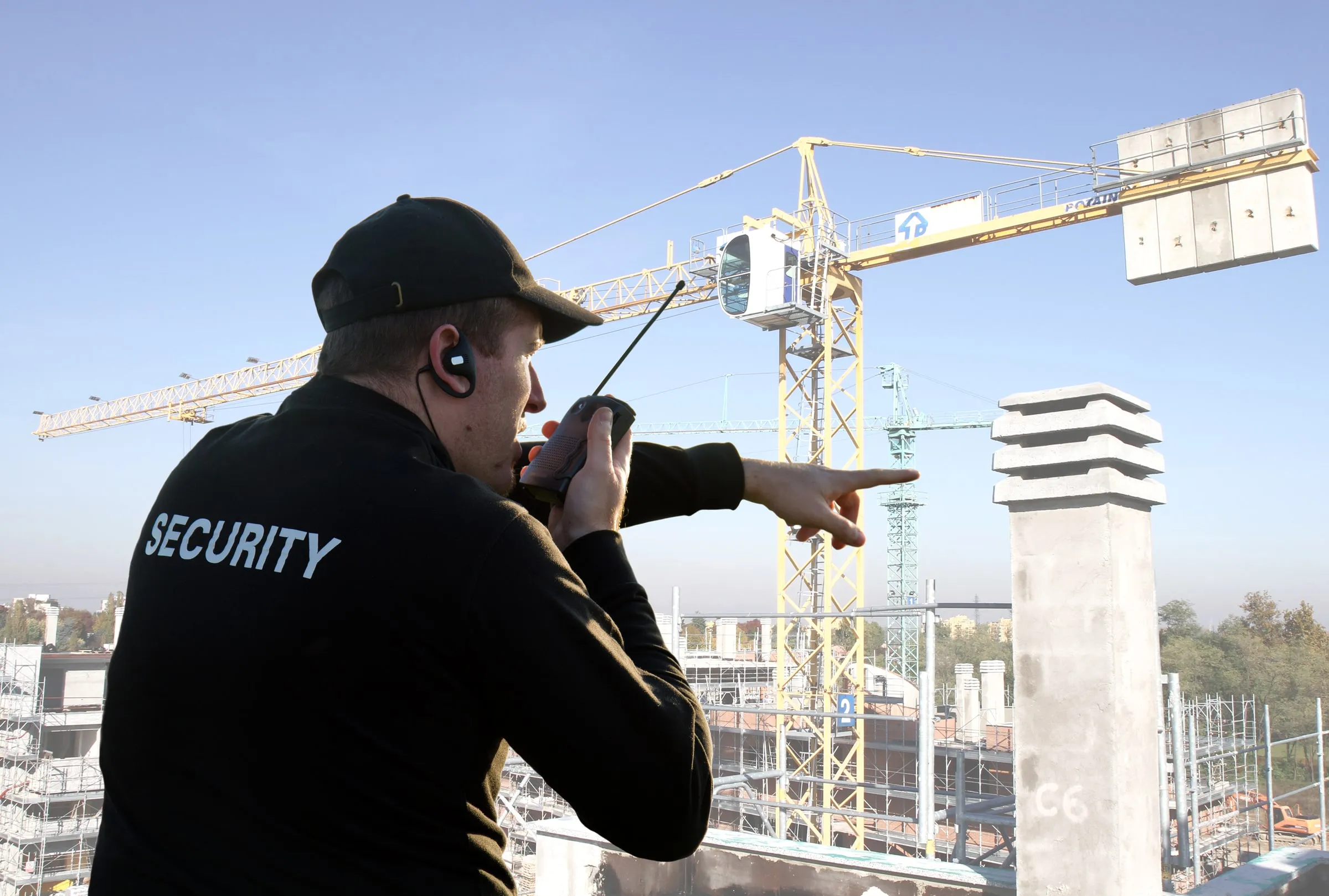 Security officer in a black SECURITY shirt speaking into a radio and pointing across a construction site with a tower crane