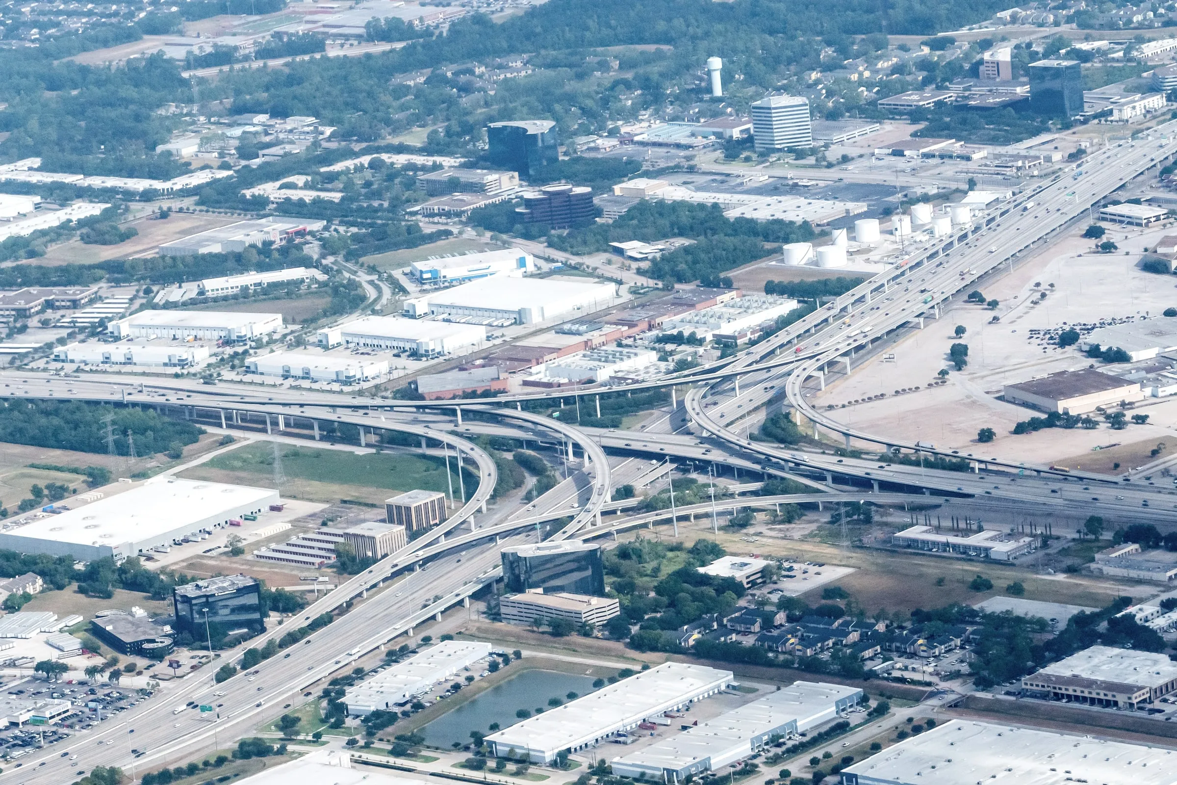 Aerial view of warehouses and an industrial park beside a freeway interchange with a rail line and tree cover