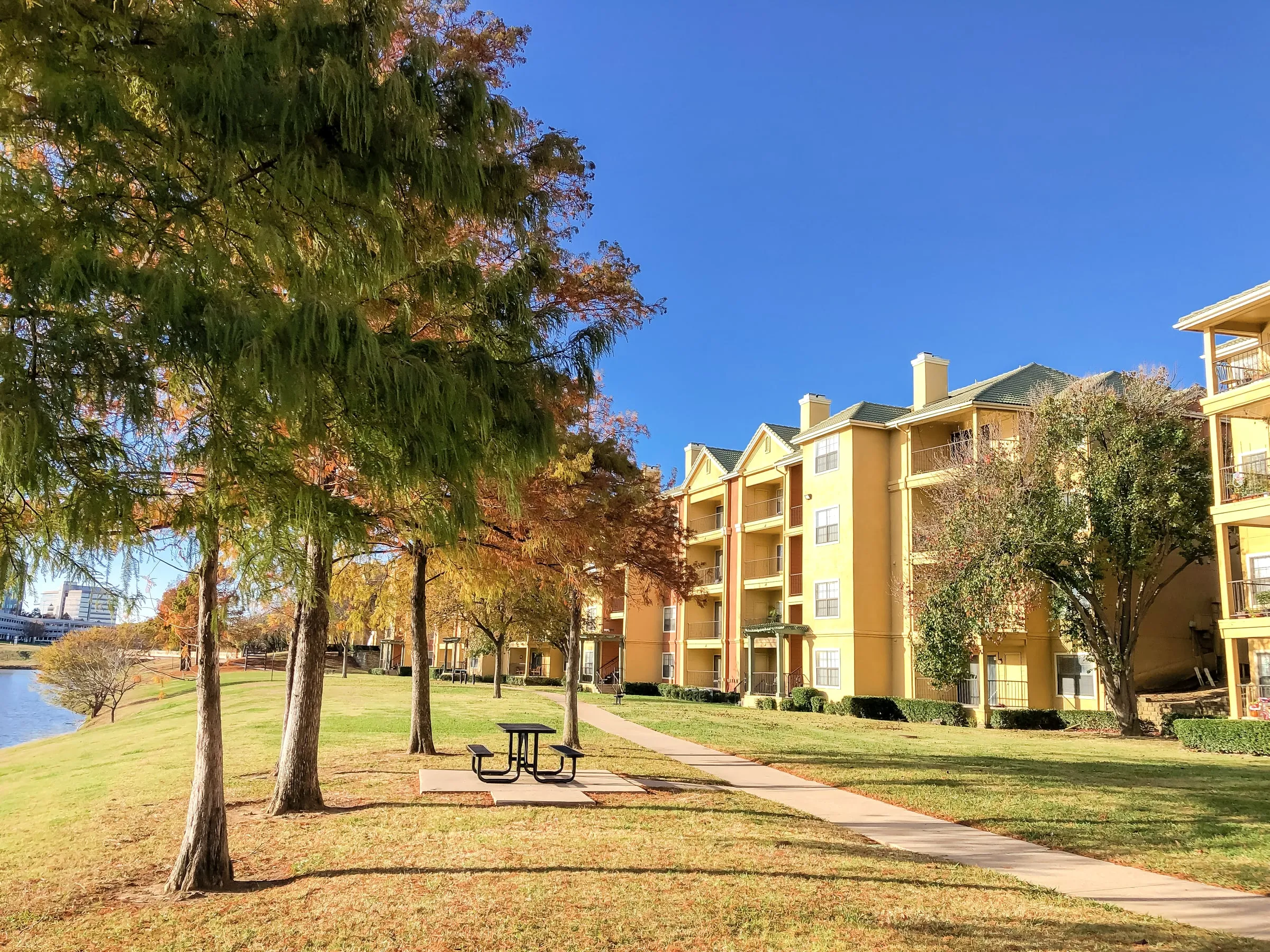 Three-storey apartment buildings beside a pond with trees and a picnic table in autumn light