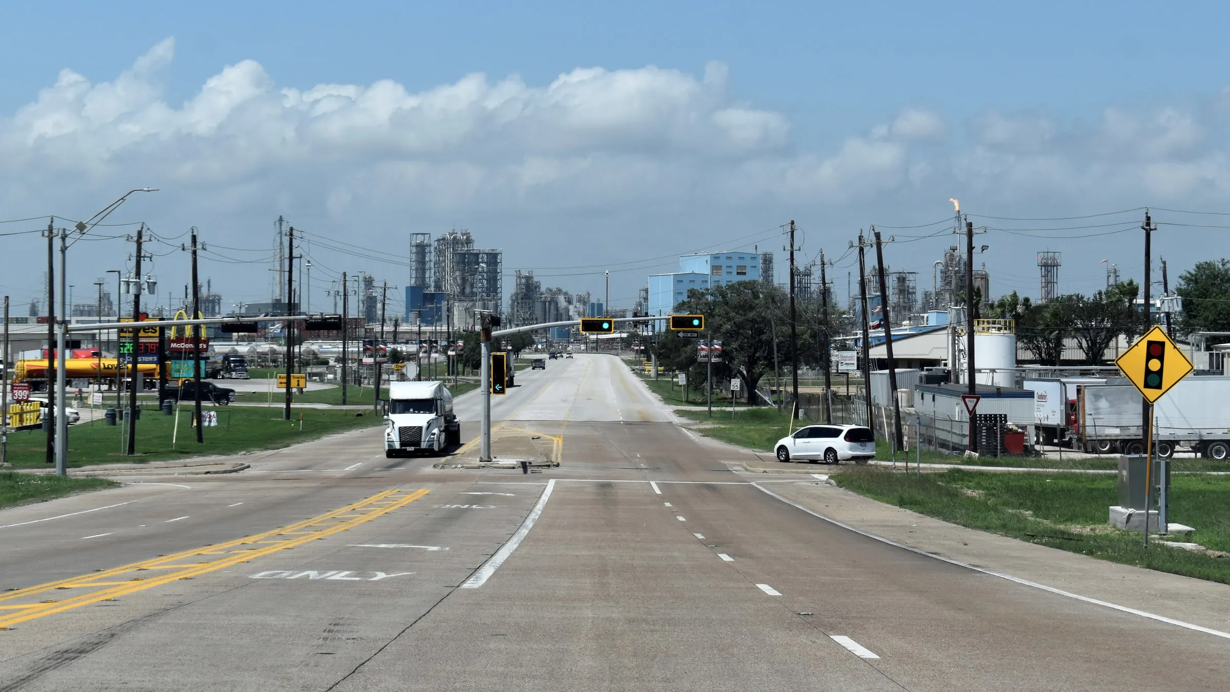Wide suburban road with a truck and cars under a clear sky, low buildings and utility poles along both sides