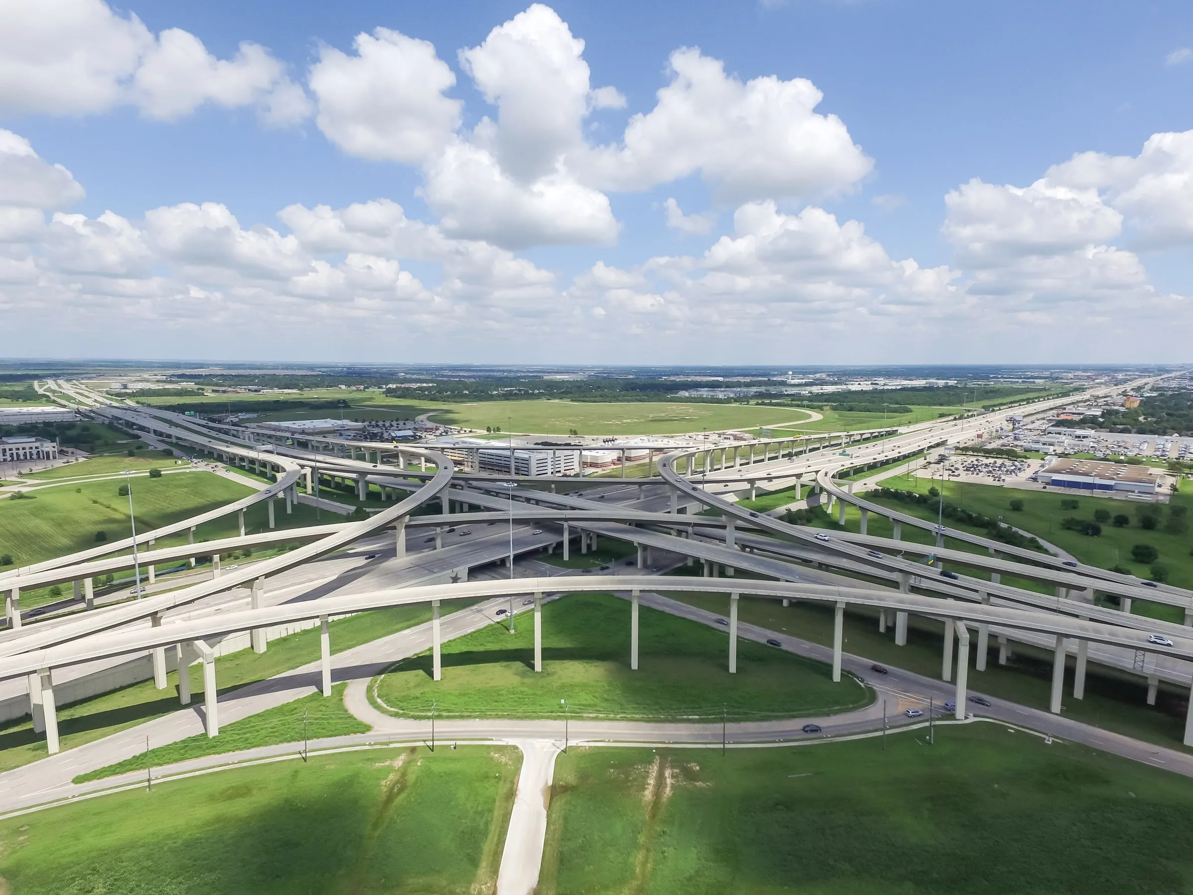 Aerial view of the Grand Parkway and Katy Freeway stack interchange under a partly cloudy sky, with flat green land and retail rooftops beyond