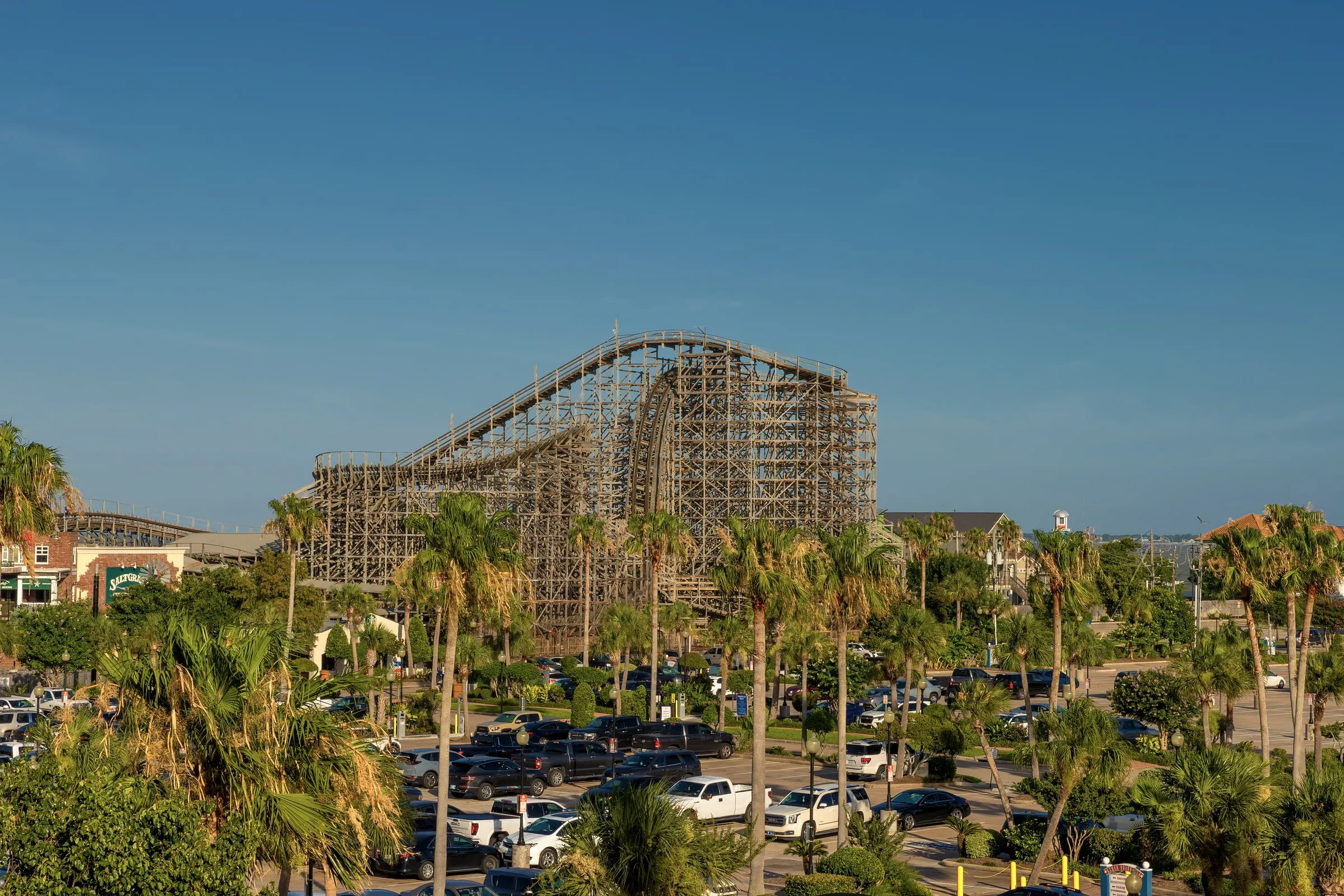 Kemah Boardwalk with a wooden roller coaster, palm trees and a full parking lot on a sunny day
