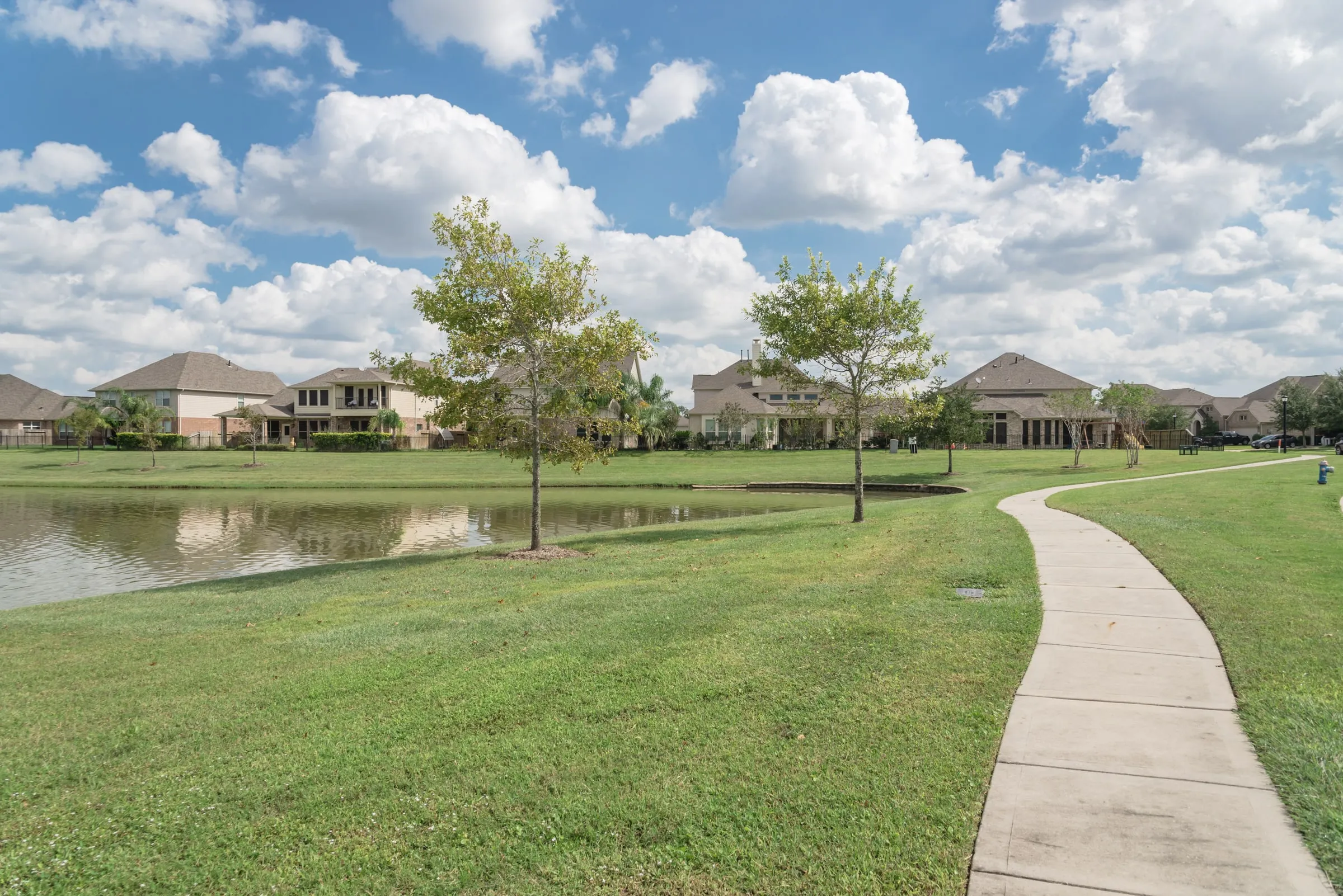 Neighborhood park with a pond, a curving path, mature trees and homes beyond under a blue sky