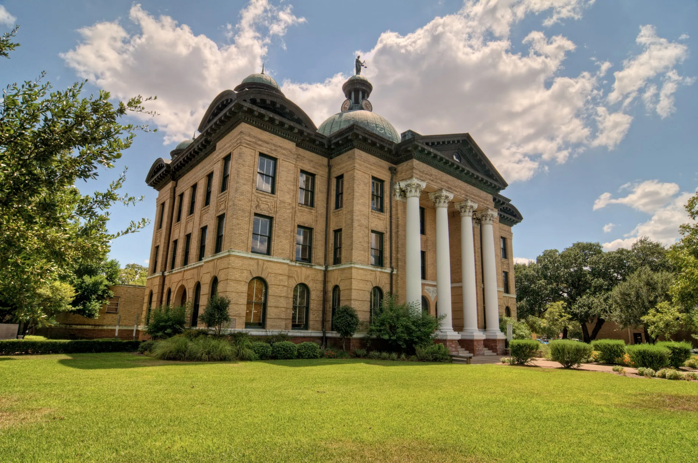 The historic Fort Bend County courthouse in Richmond, yellow brick with green domes and tall white columns, under a blue sky with cumulus clouds