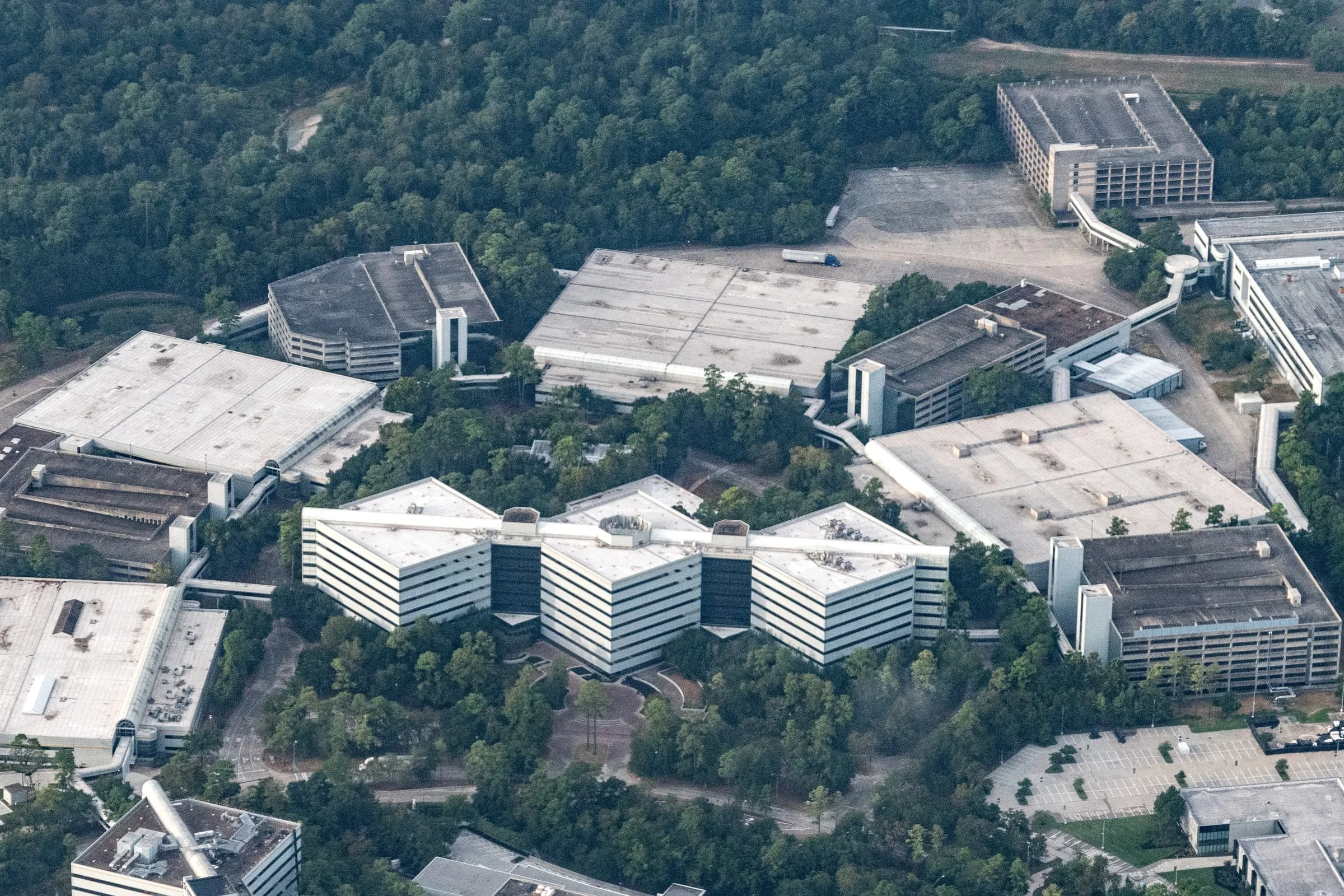 Aerial view of a wooded office and warehouse campus with flat white roofs, parking garages, a large empty surface lot and a single semi-trailer