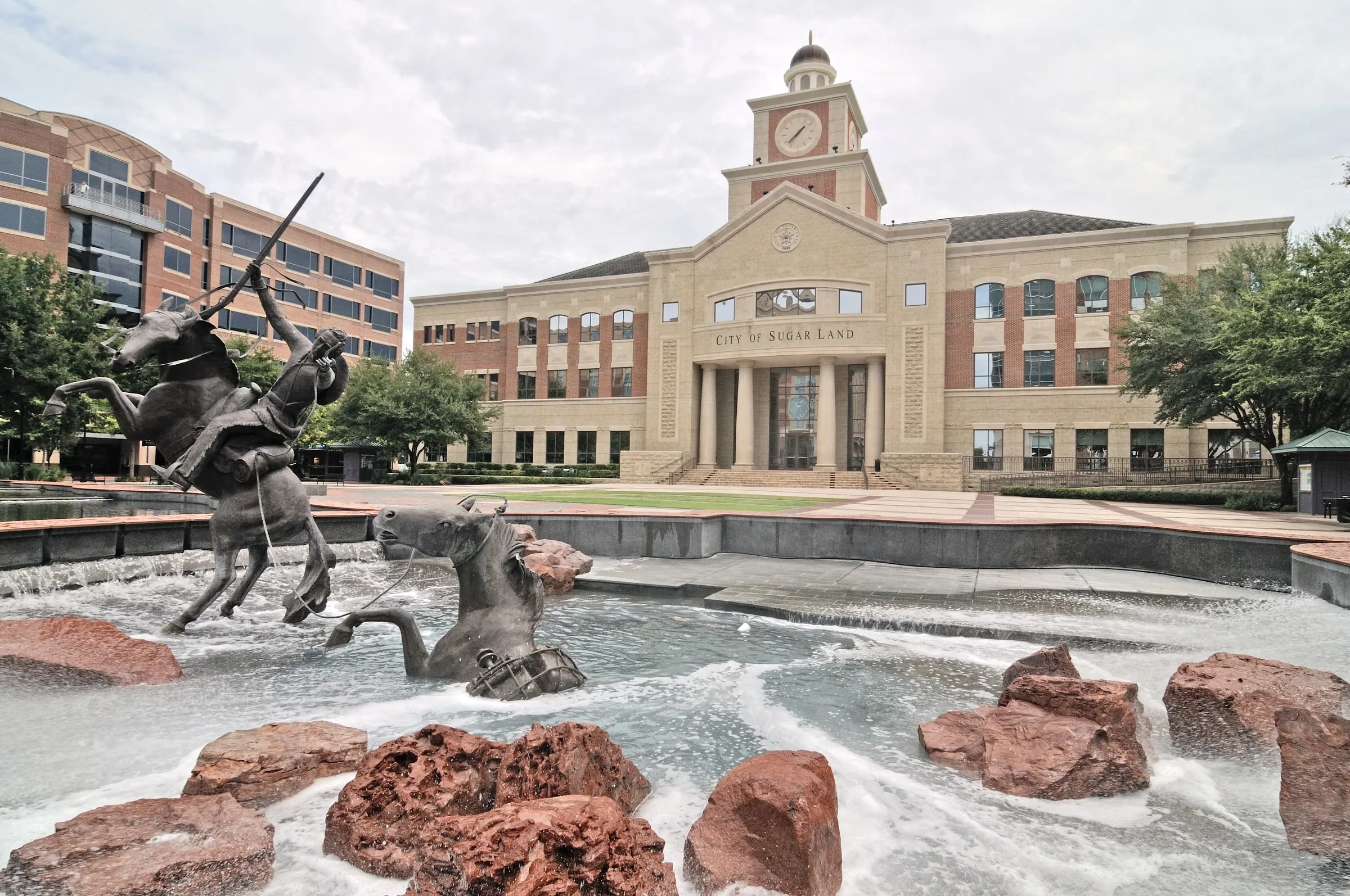 Sugar Land City Hall at Town Square, with its clock tower and columned entrance behind a fountain and bronze horse-and-rider sculpture on an overcast day