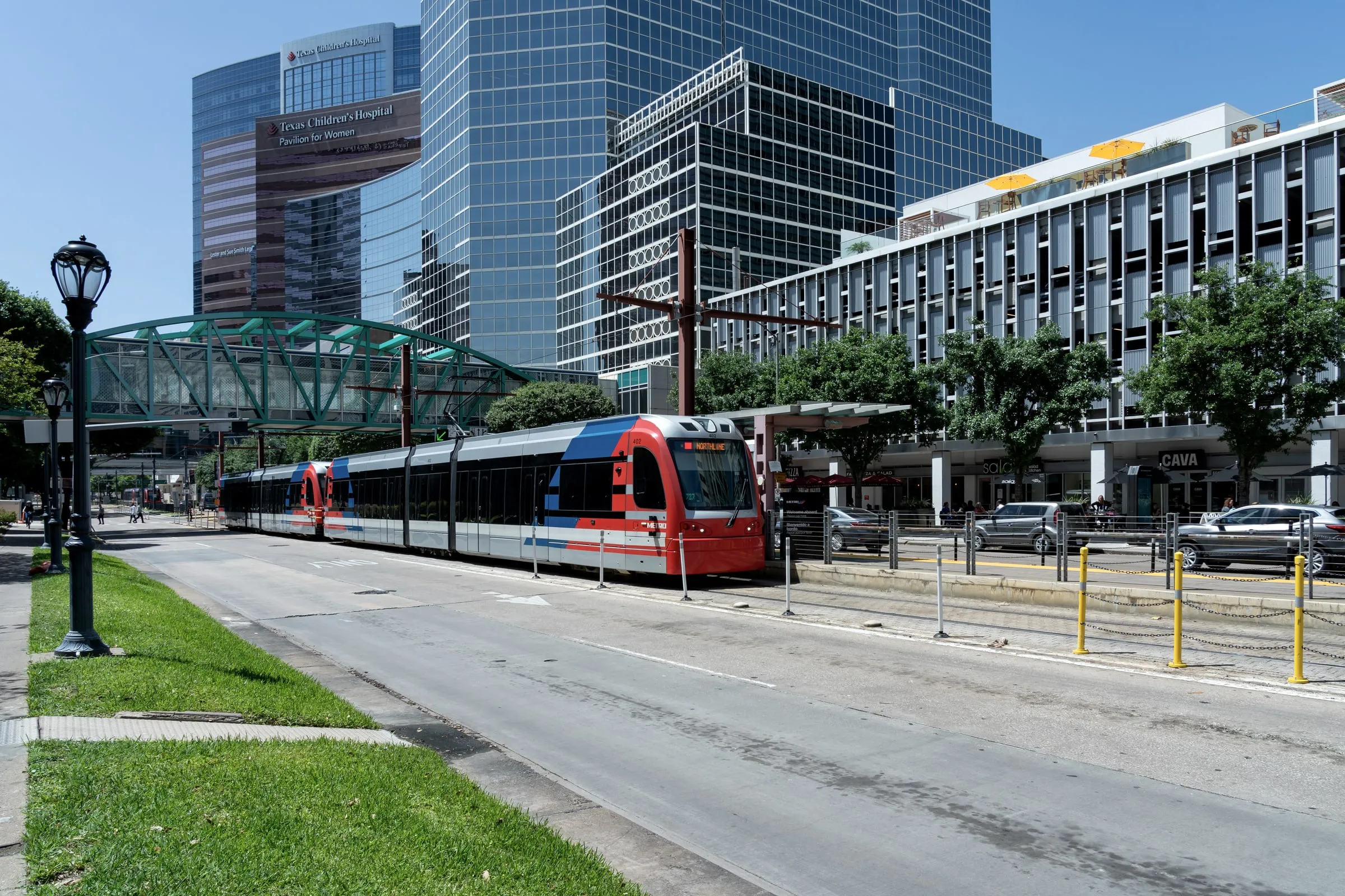 Red METRORail light rail train on a tree-lined street between glass medical office buildings