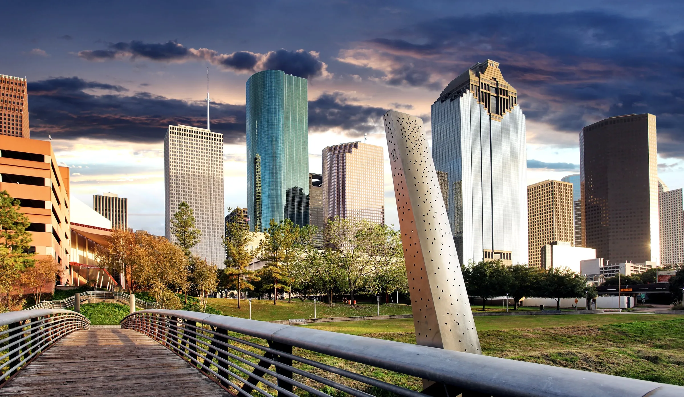 Downtown Houston skyline seen from a pedestrian bridge with a tall white sculpture, under scattered clouds