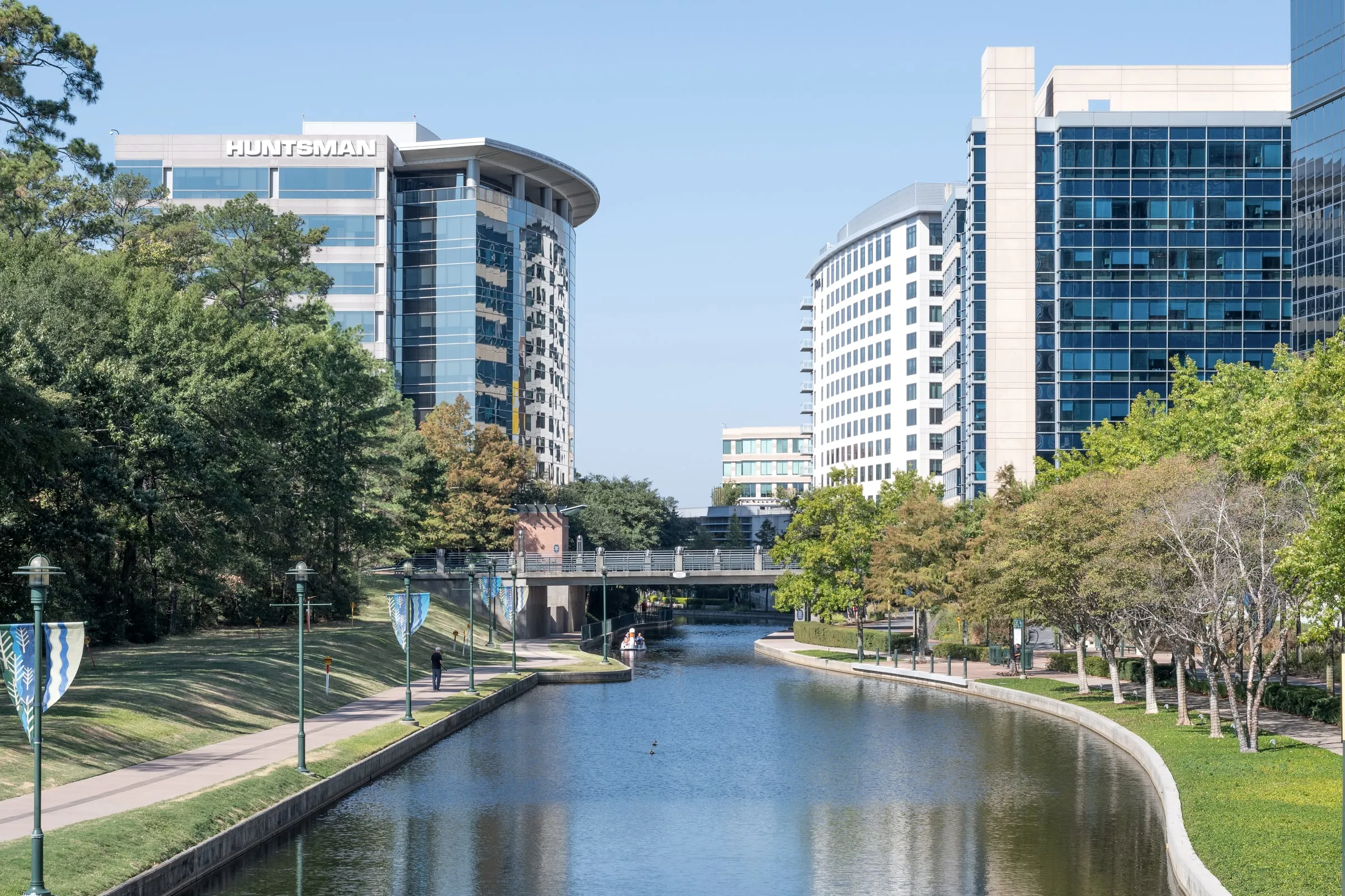 The Woodlands Waterway lined with trees, walkways and glass office buildings under a blue sky
