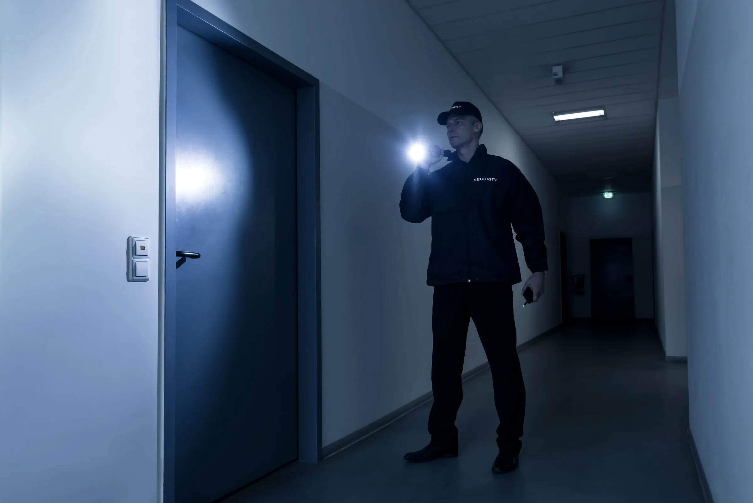 Security officer in a dark jacket lettered SECURITY and a cap shines a flashlight on a closed door in a dim office corridor