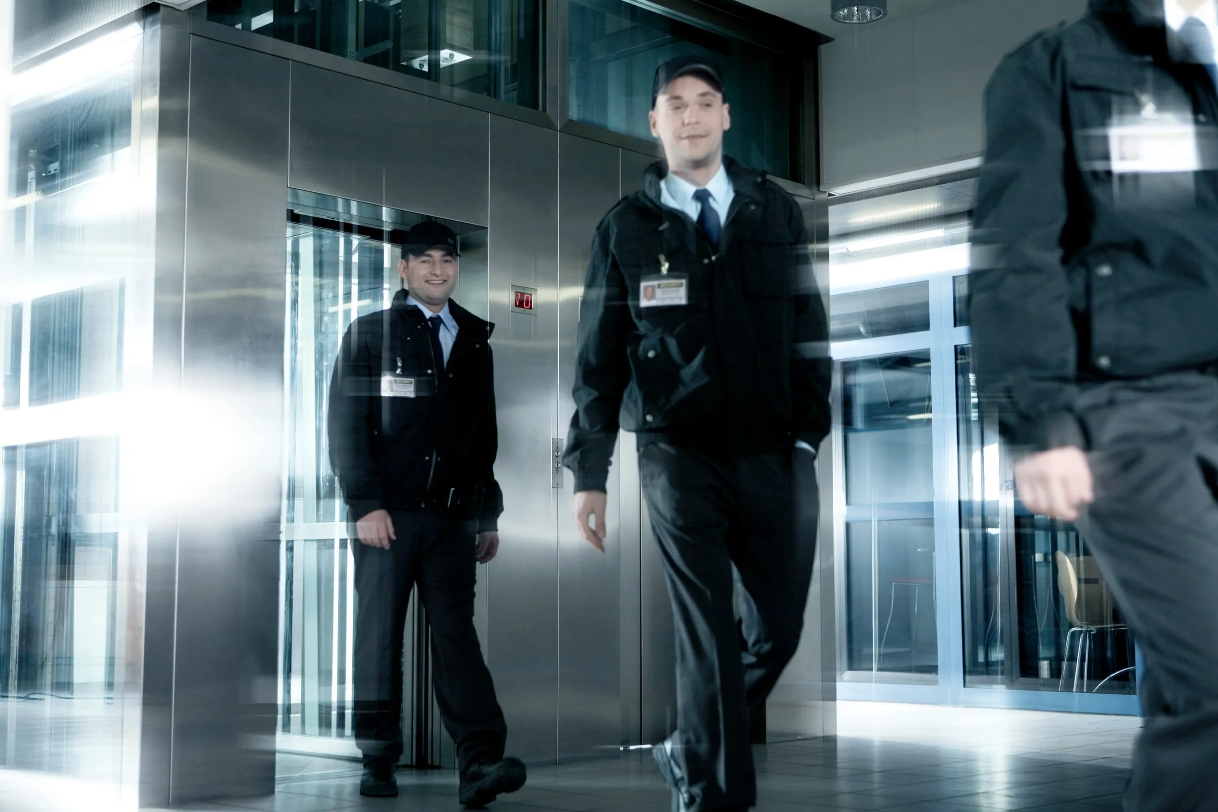 Three security officers in dark jackets, caps and ID badges walk through a bright lobby past stainless-steel elevator doors