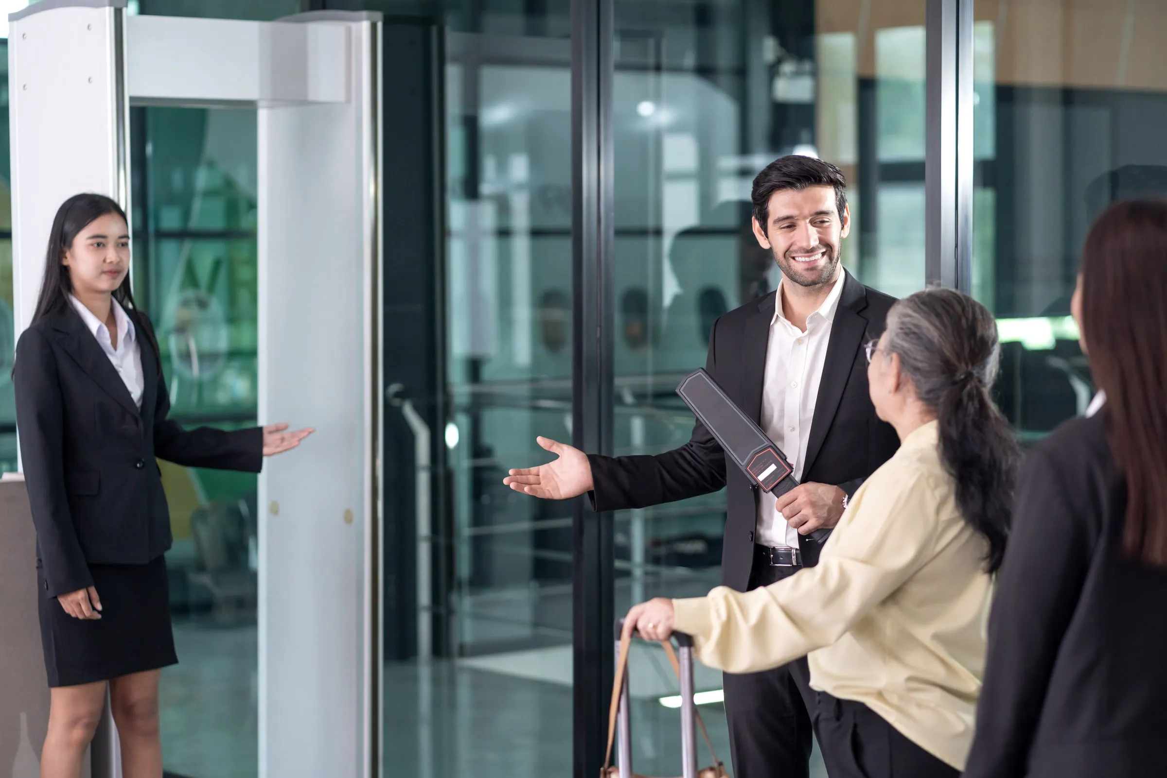 A suited officer holding a handheld metal-detector wand welcomes two visitors toward a walk-through screening arch in a glass-walled lobby