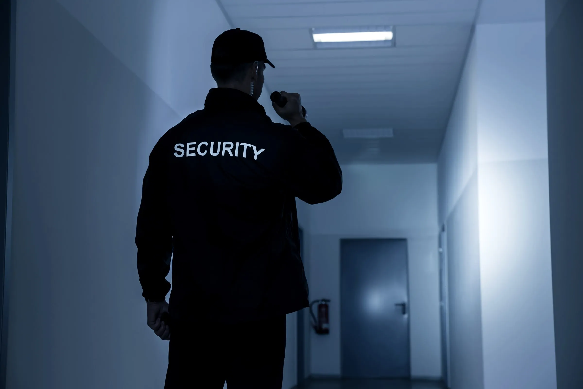 Officer in a dark jacket lettered SECURITY raises a flashlight in a dim corridor with a fire extinguisher and a closed door at the far end