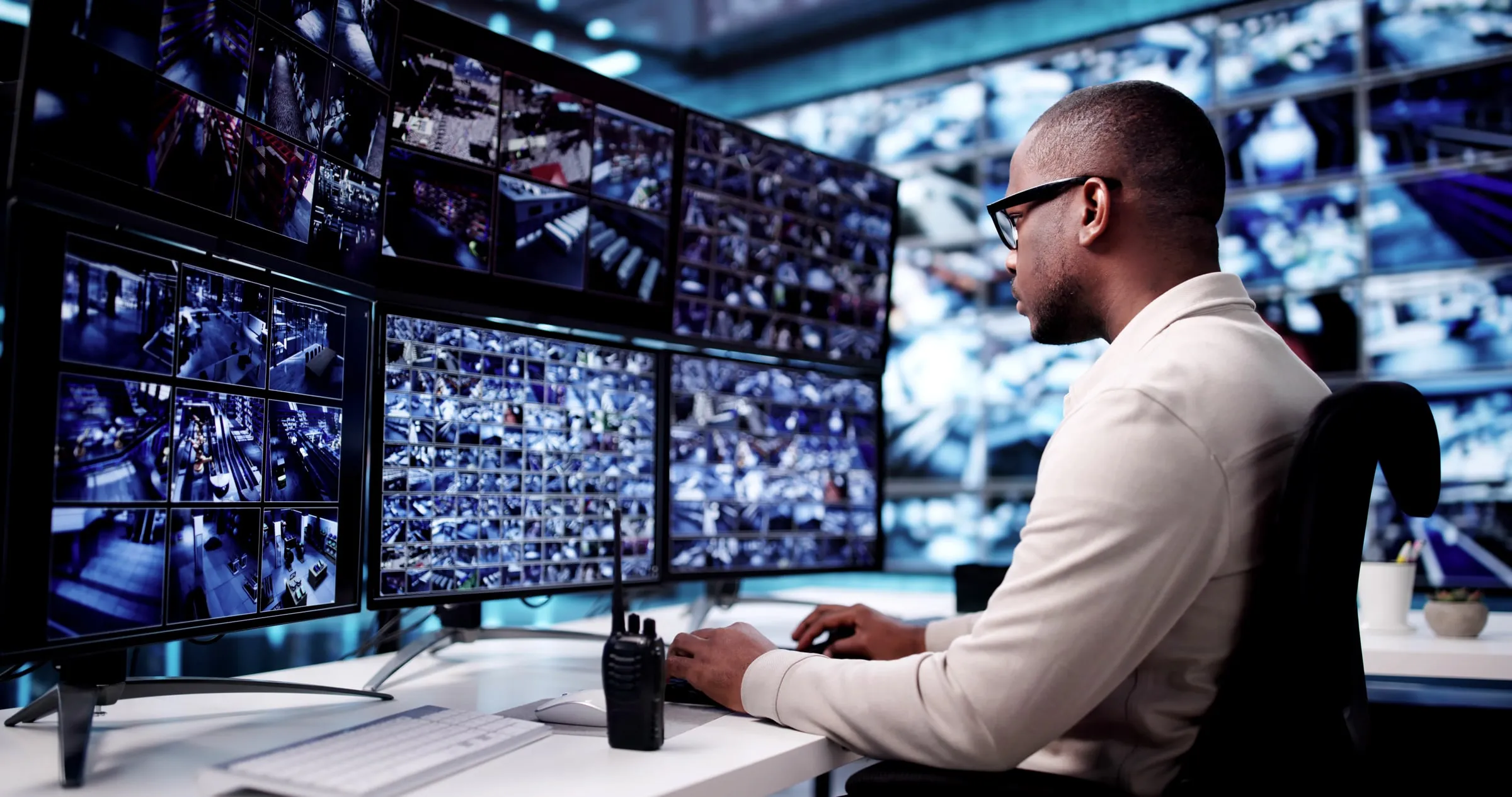 A seated operator in glasses and a light shirt watches a bank of monitors filled with camera feeds, a handheld radio beside the keyboard