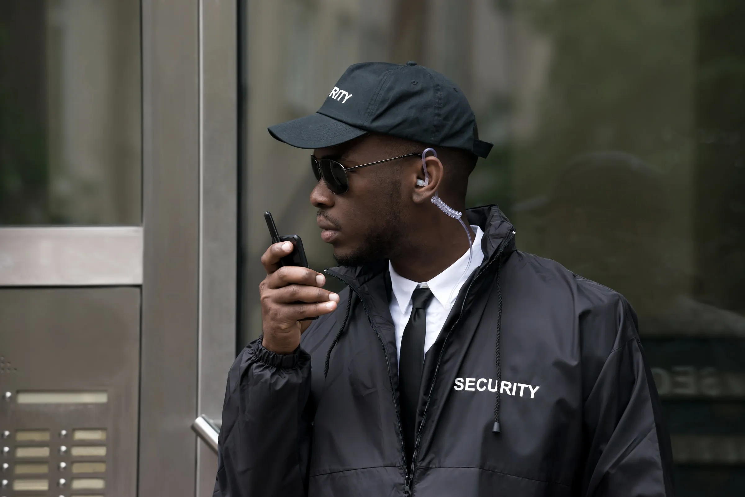 Security officer in a black SECURITY jacket and cap speaking into a handheld radio outside a glass entrance door