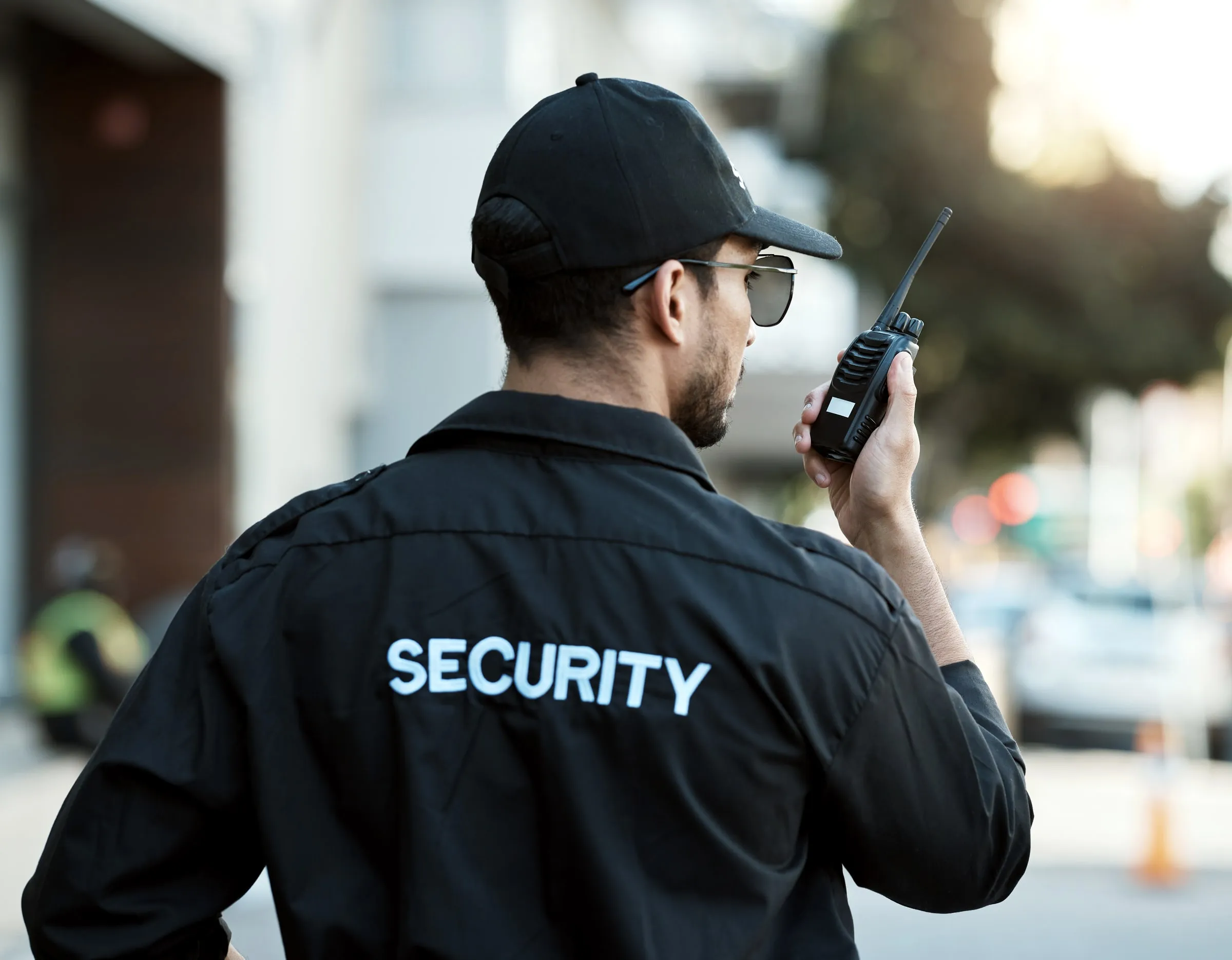 Rear view of a security officer in a black SECURITY shirt and cap talking on a radio on a street with blurred traffic behind
