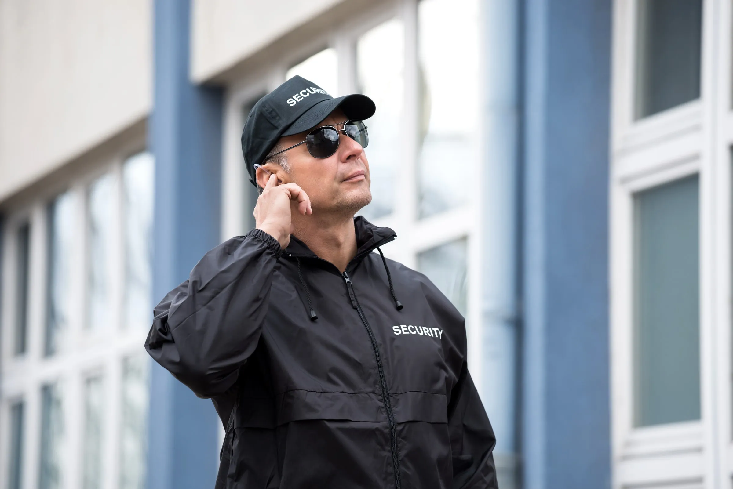 Security officer in a black SECURITY cap, sunglasses and jacket touching his earpiece outside a building with blue columns and tall windows