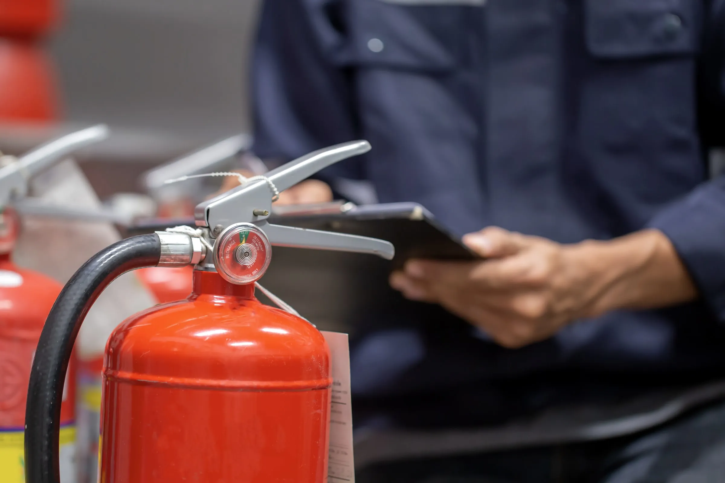 Red fire extinguisher with a pressure gauge and black hose in sharp focus, a person in a navy work shirt holding a clipboard behind it
