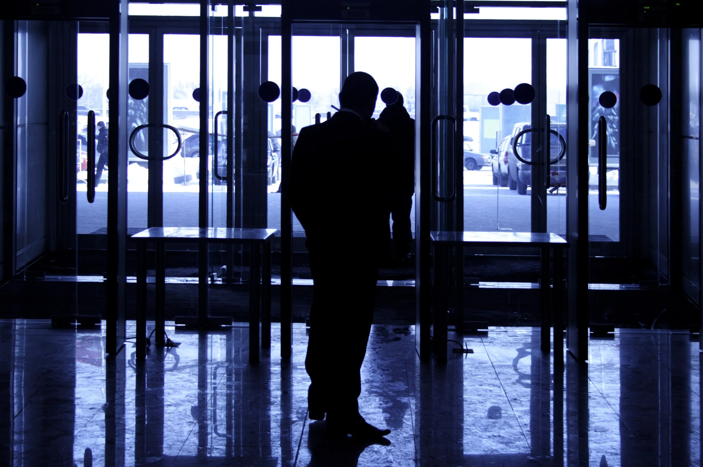 Silhouettes of two people at a glass entrance of a building lobby, seen against bright daylight