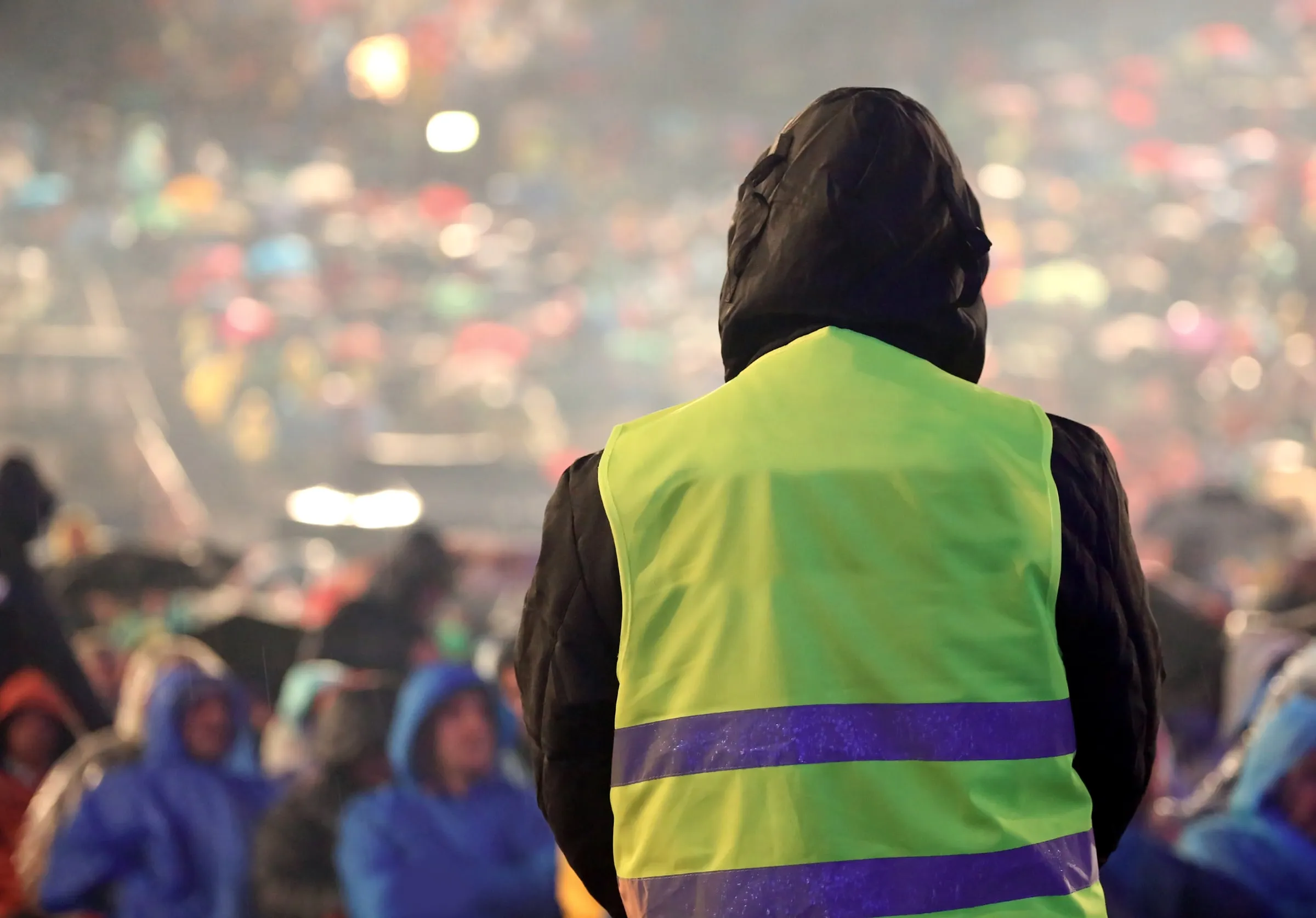 Officer in a yellow high-visibility vest and hooded black jacket, seen from behind, facing a large crowd in rain jackets at a night event