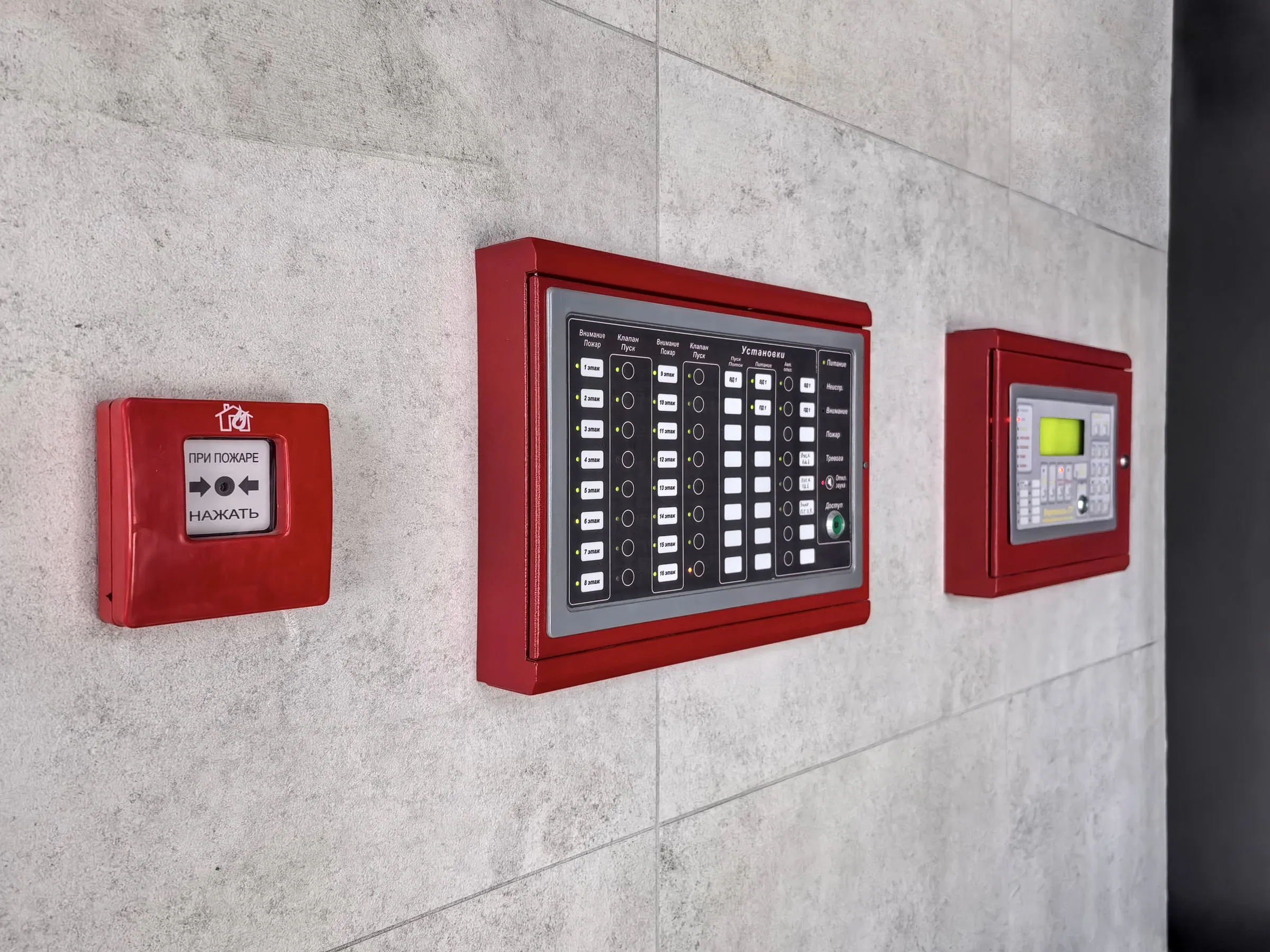 Red manual pull station and two red fire alarm control panels mounted in a row on a grey stone-tiled wall