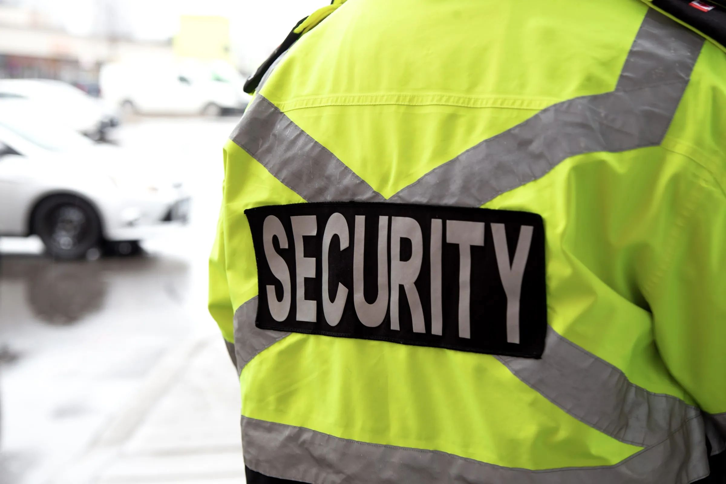 Back of a security officer in a hi-vis yellow jacket with a black SECURITY panel, in a parking lot