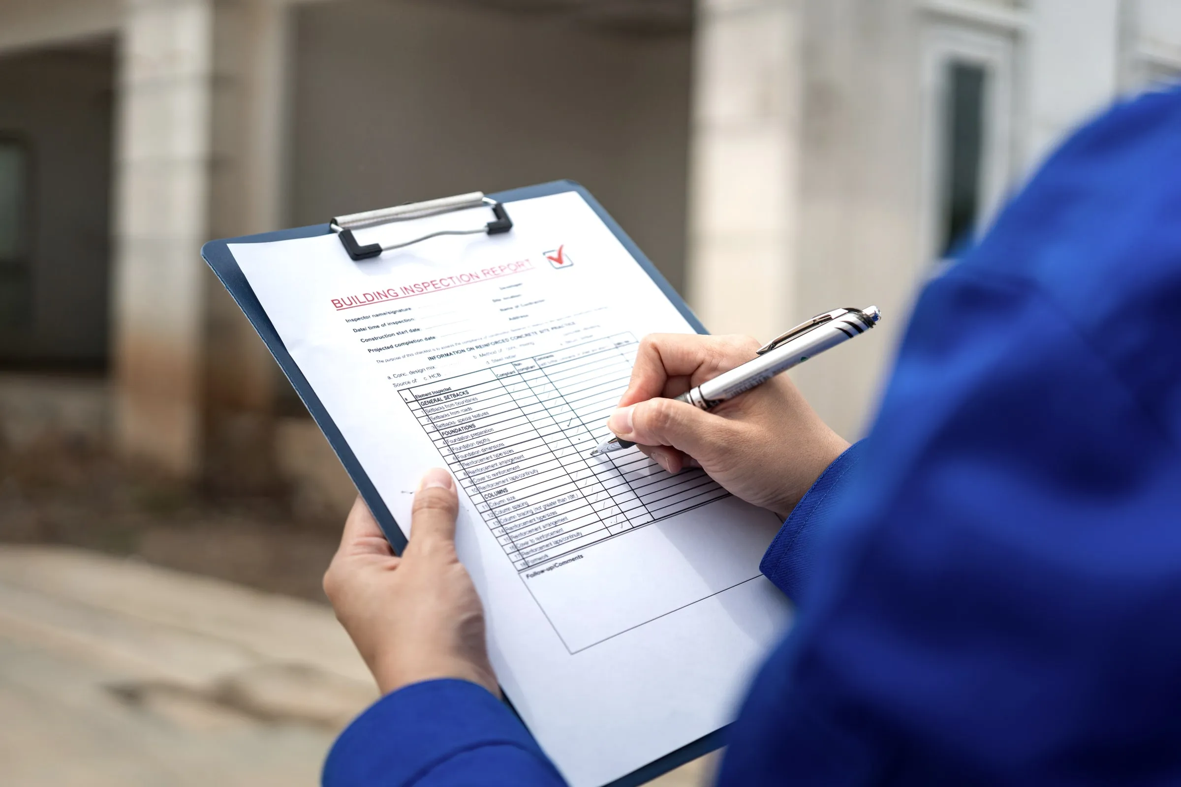 Hands in a blue jacket holding a clipboard with a building inspection checklist and a pen