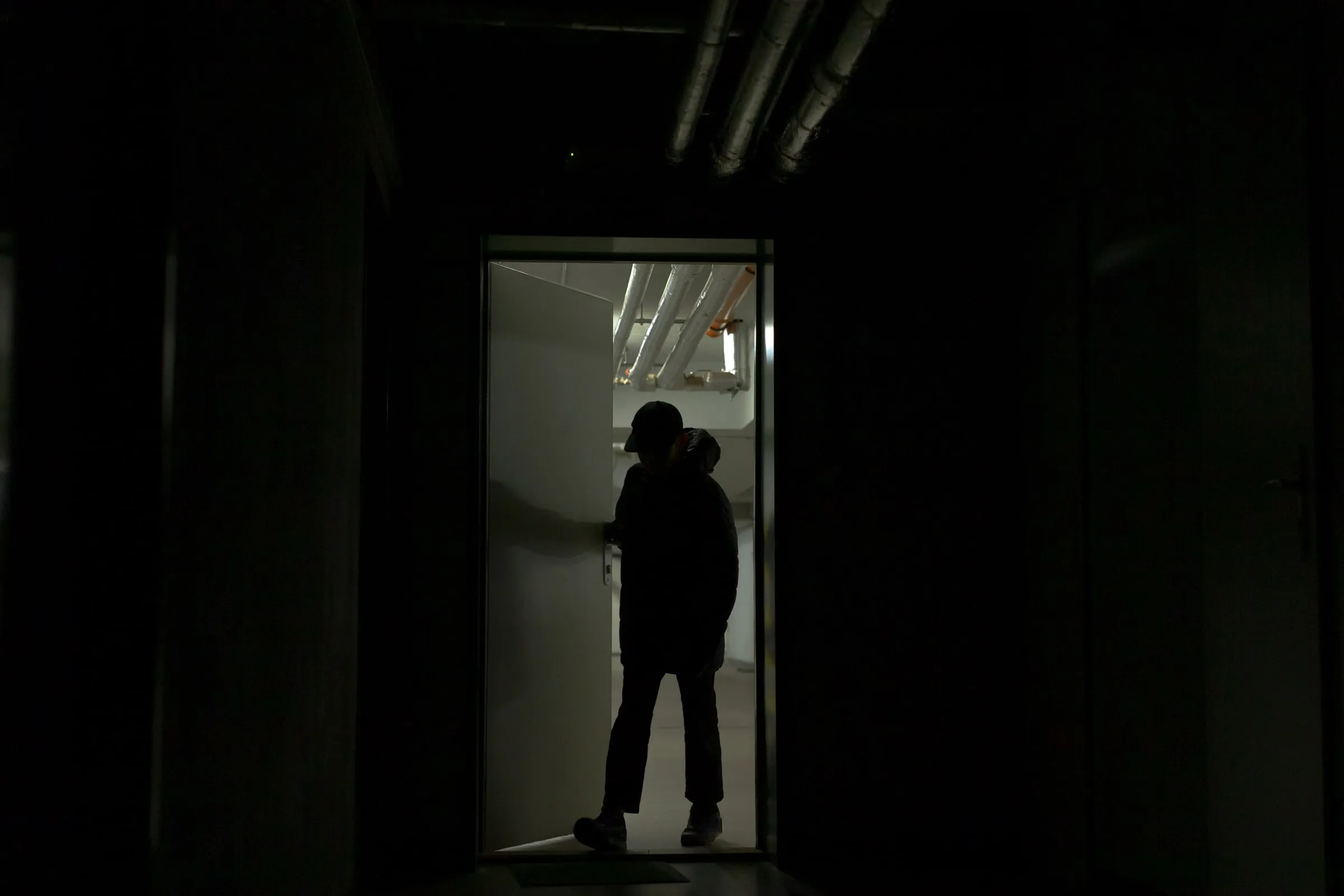 Silhouetted person in a cap at the open door of a lit mechanical room, insulated pipes running along the ceiling beyond