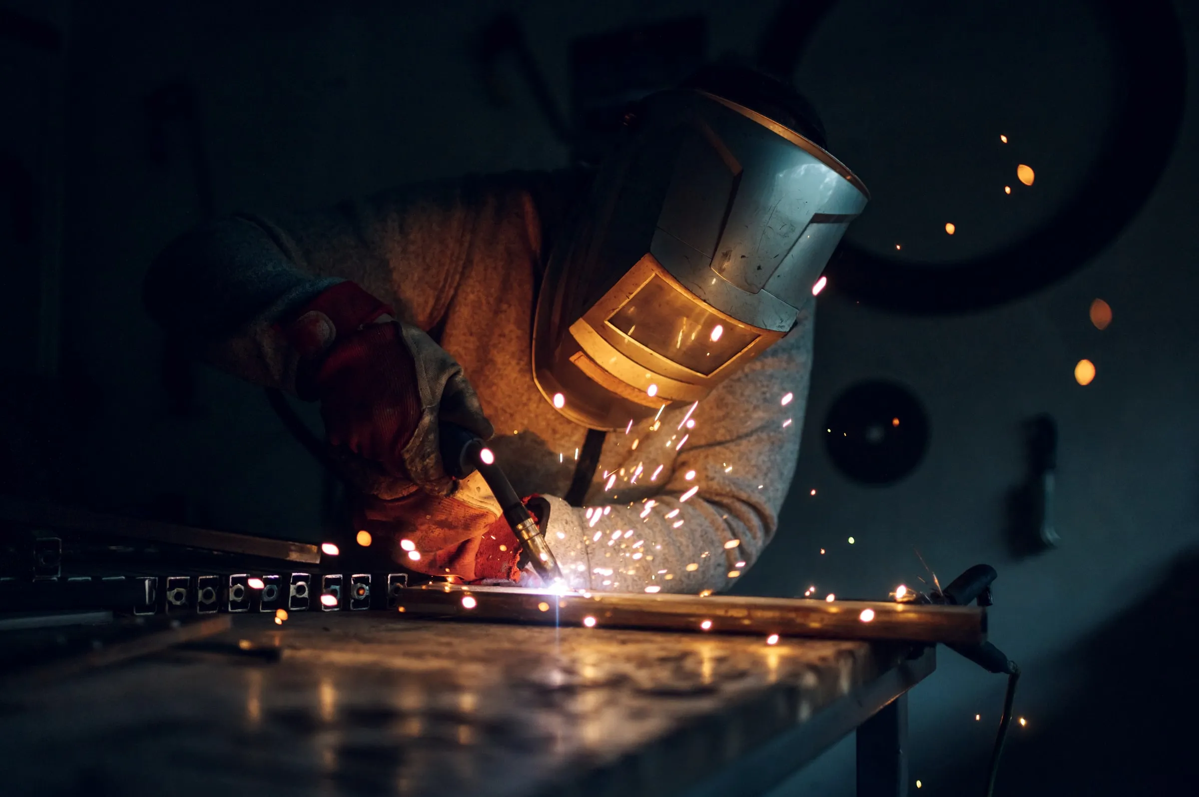 Welder in a flip-down helmet and red gloves running a bead on a steel frame at a workbench, sparks scattering into a dark shop