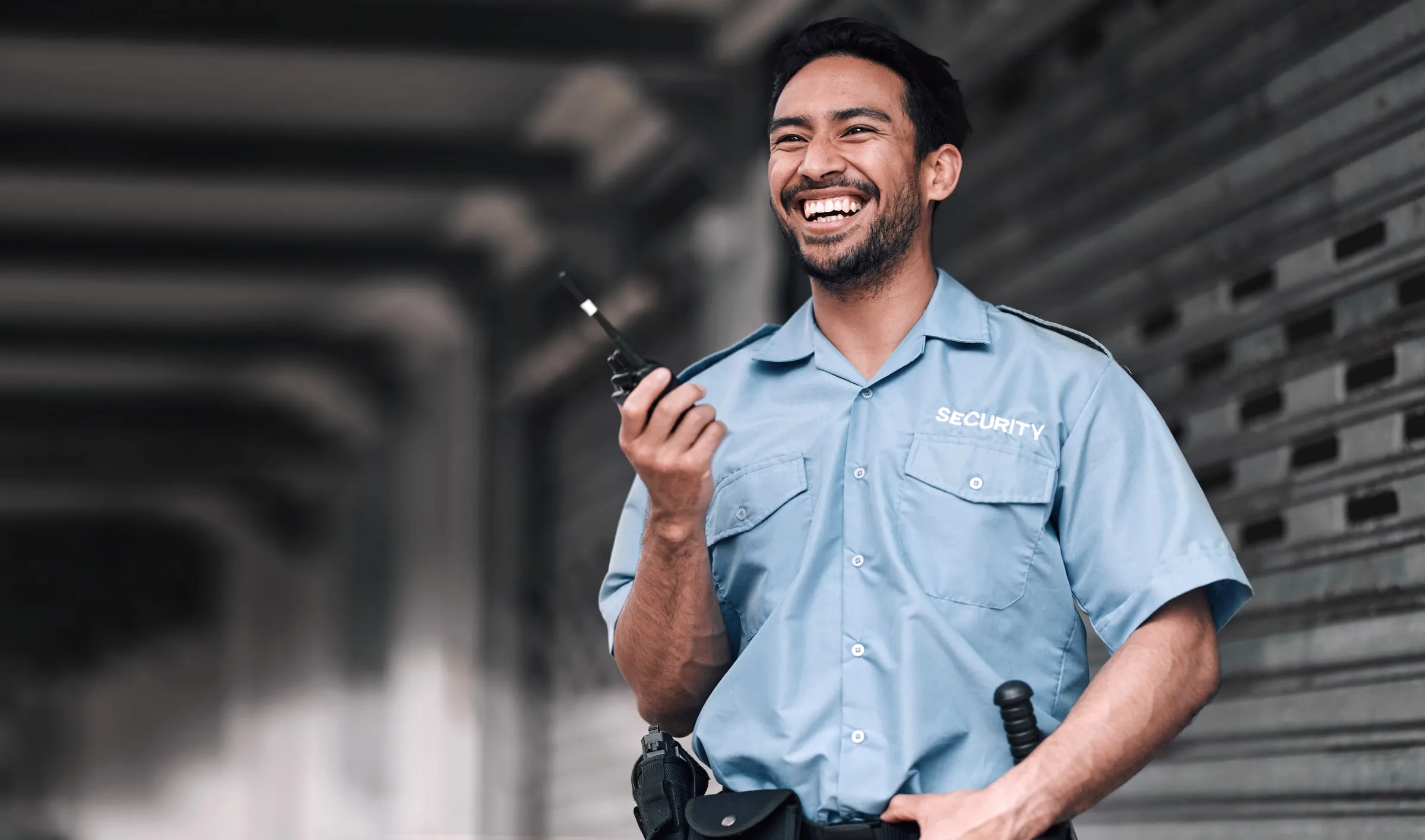 Smiling security officer in a light blue SECURITY shirt holding a handheld radio inside a concrete parking structure