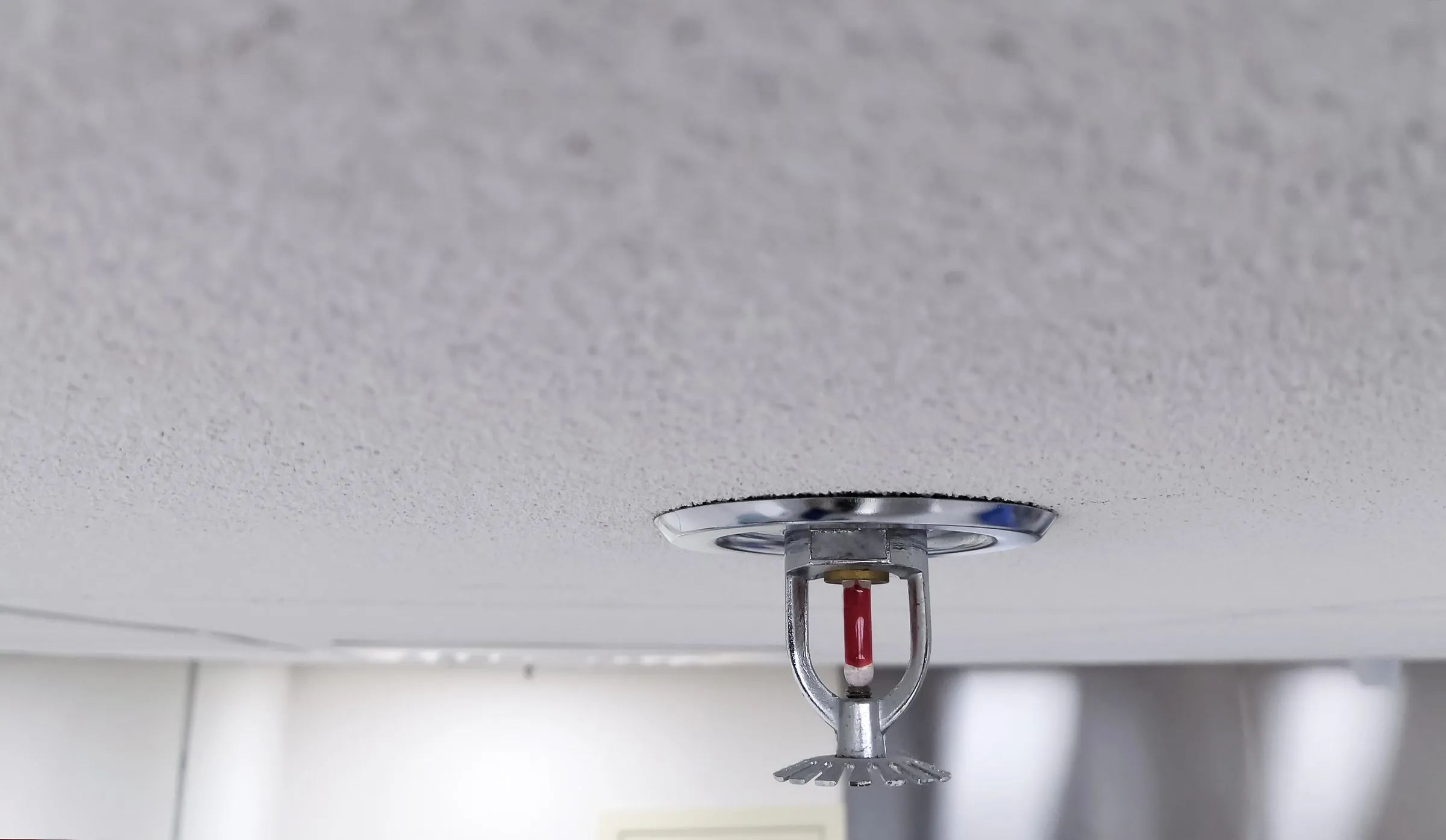 Close view of a chrome sprinkler head with a red glass bulb hanging from a white textured ceiling