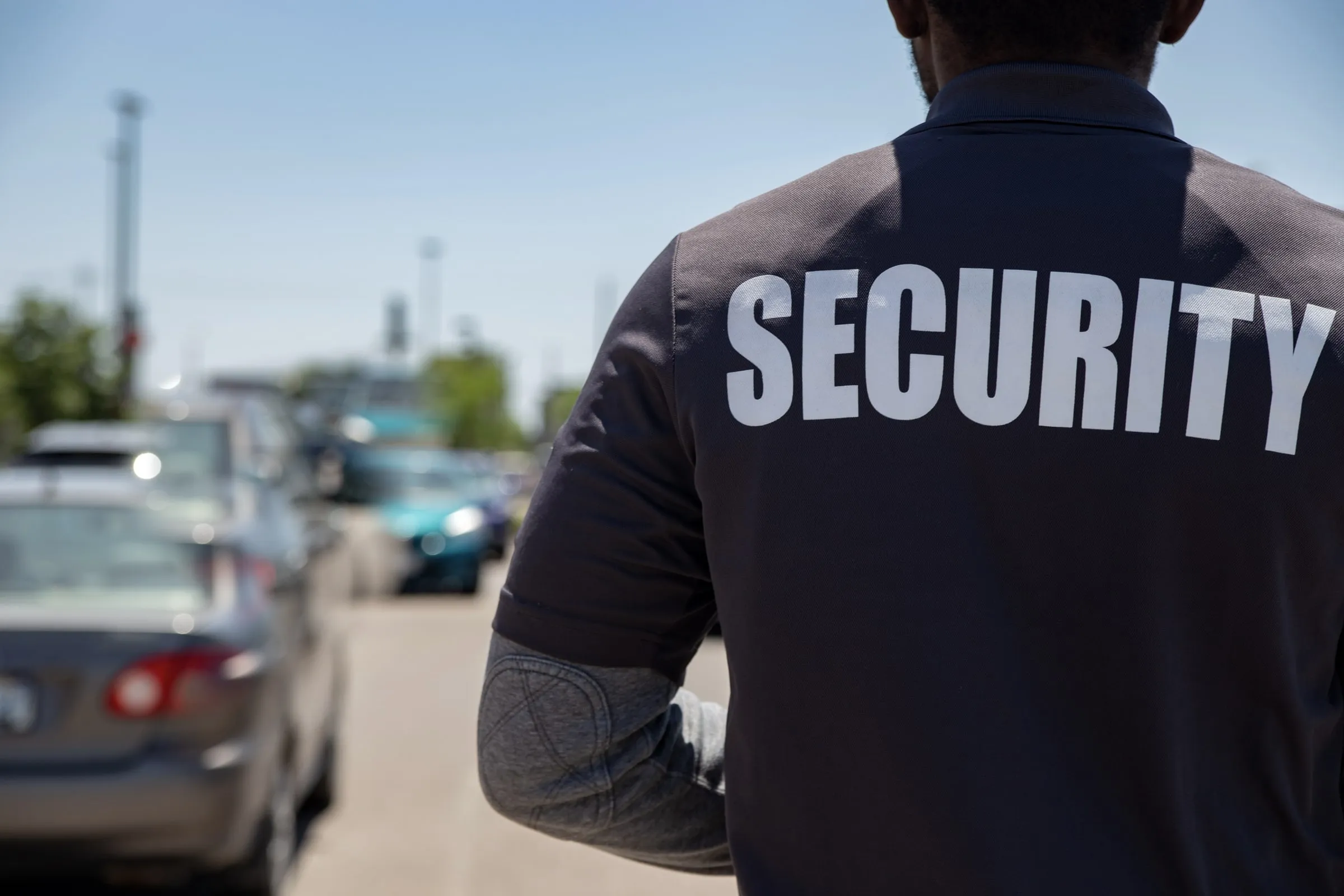Security officer seen from behind in a dark polo lettered SECURITY, standing in a sunlit parking lot with cars blurred beyond