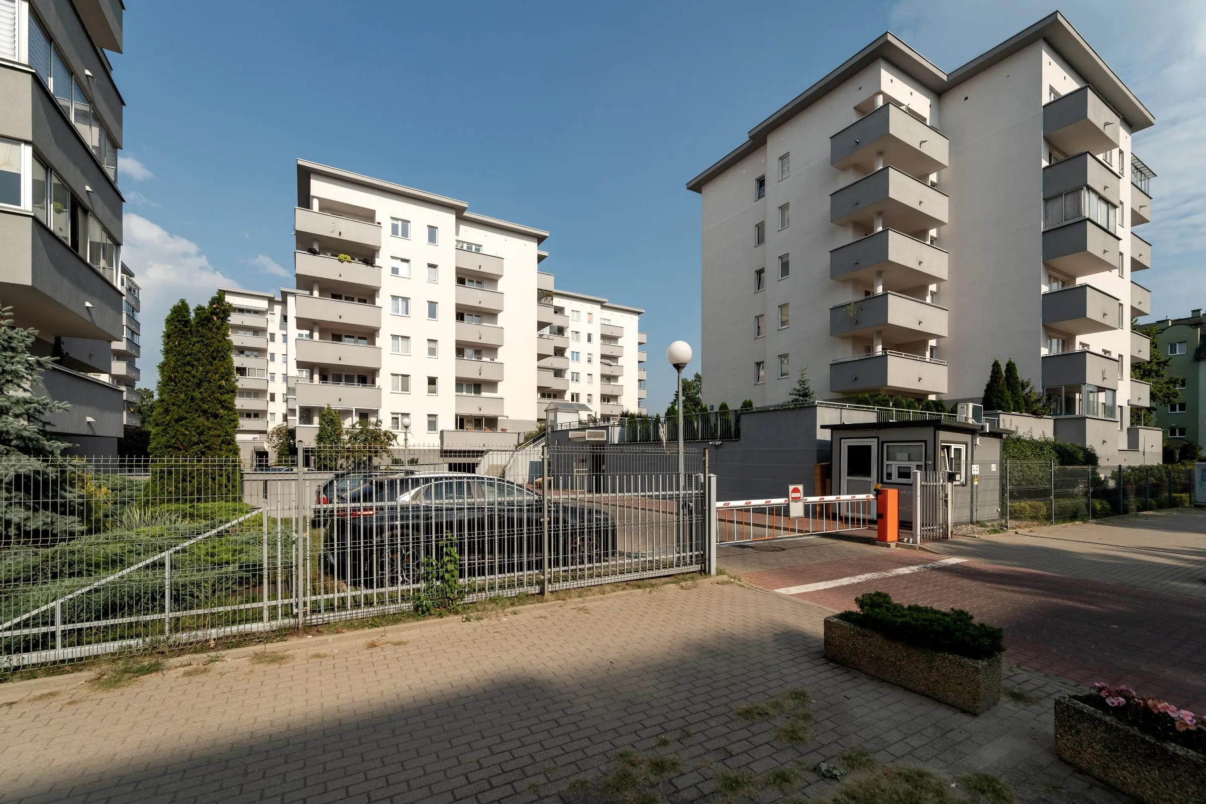 Barrier-arm vehicle entrance with a small grey guardhouse and steel fencing at a mid-rise apartment complex on a sunny day