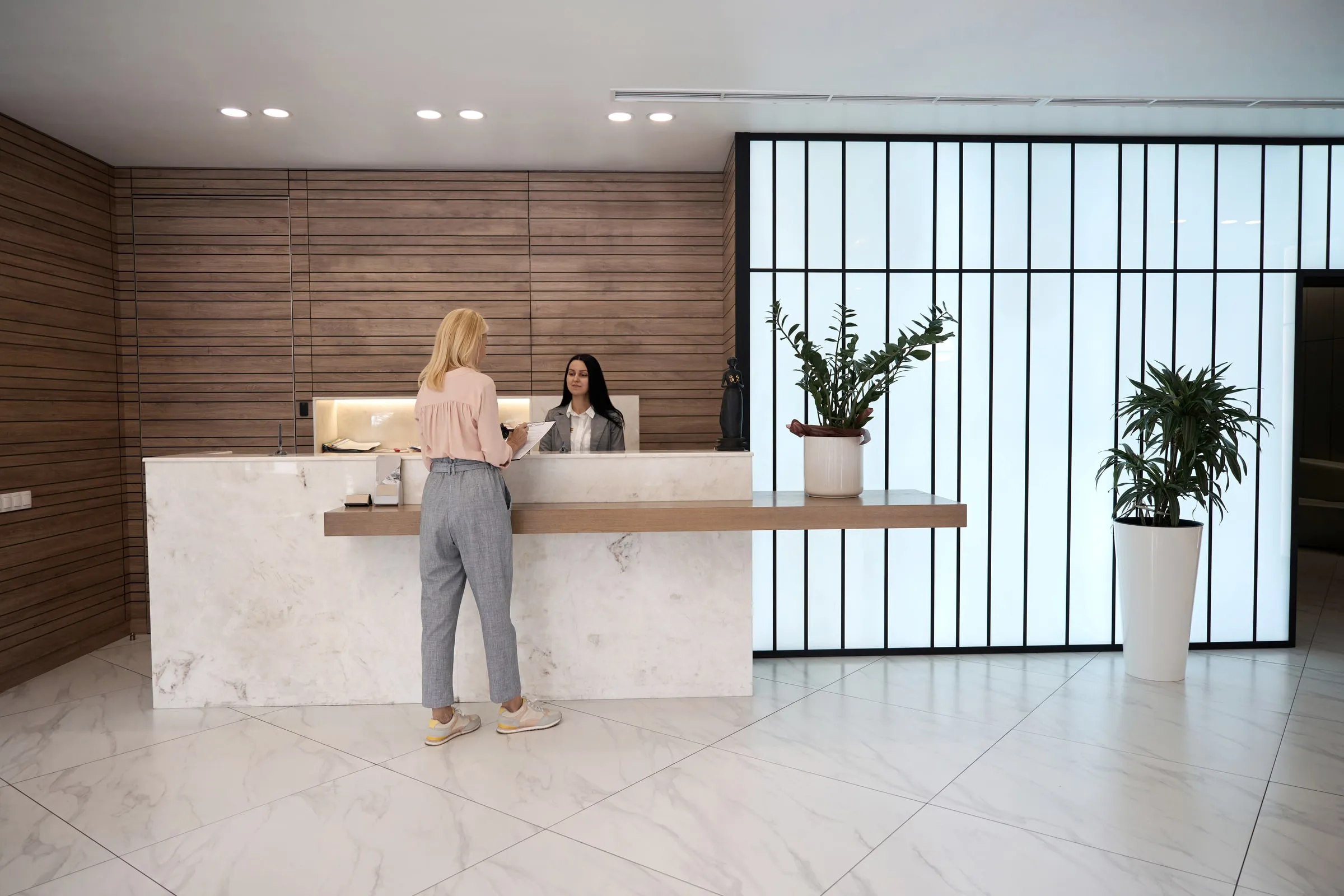 Modern office lobby with a white reception desk, wood panelling, a receptionist and a visitor being greeted