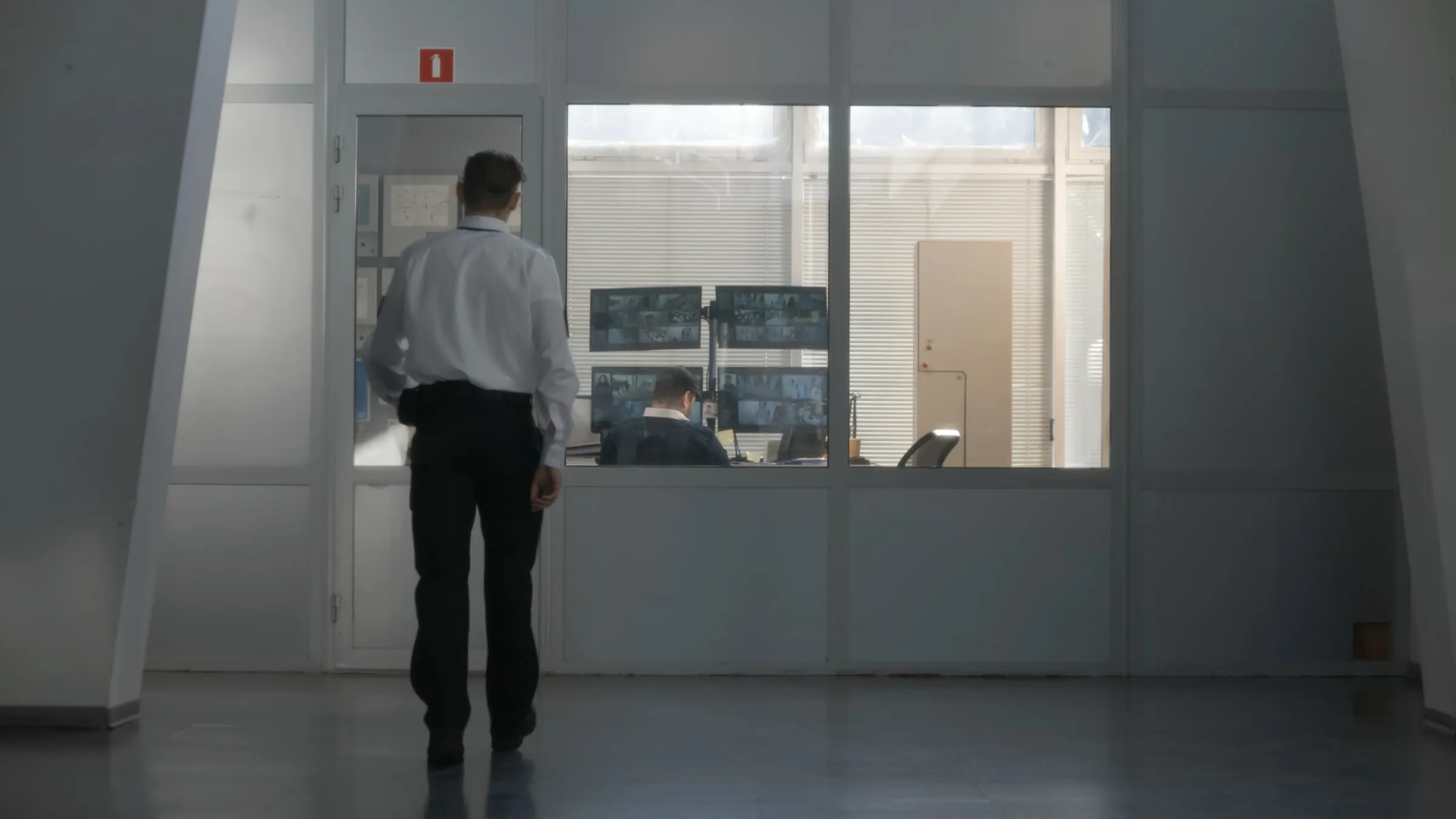 Officer in a white shirt walking toward a glass-walled security office where a colleague watches camera monitors