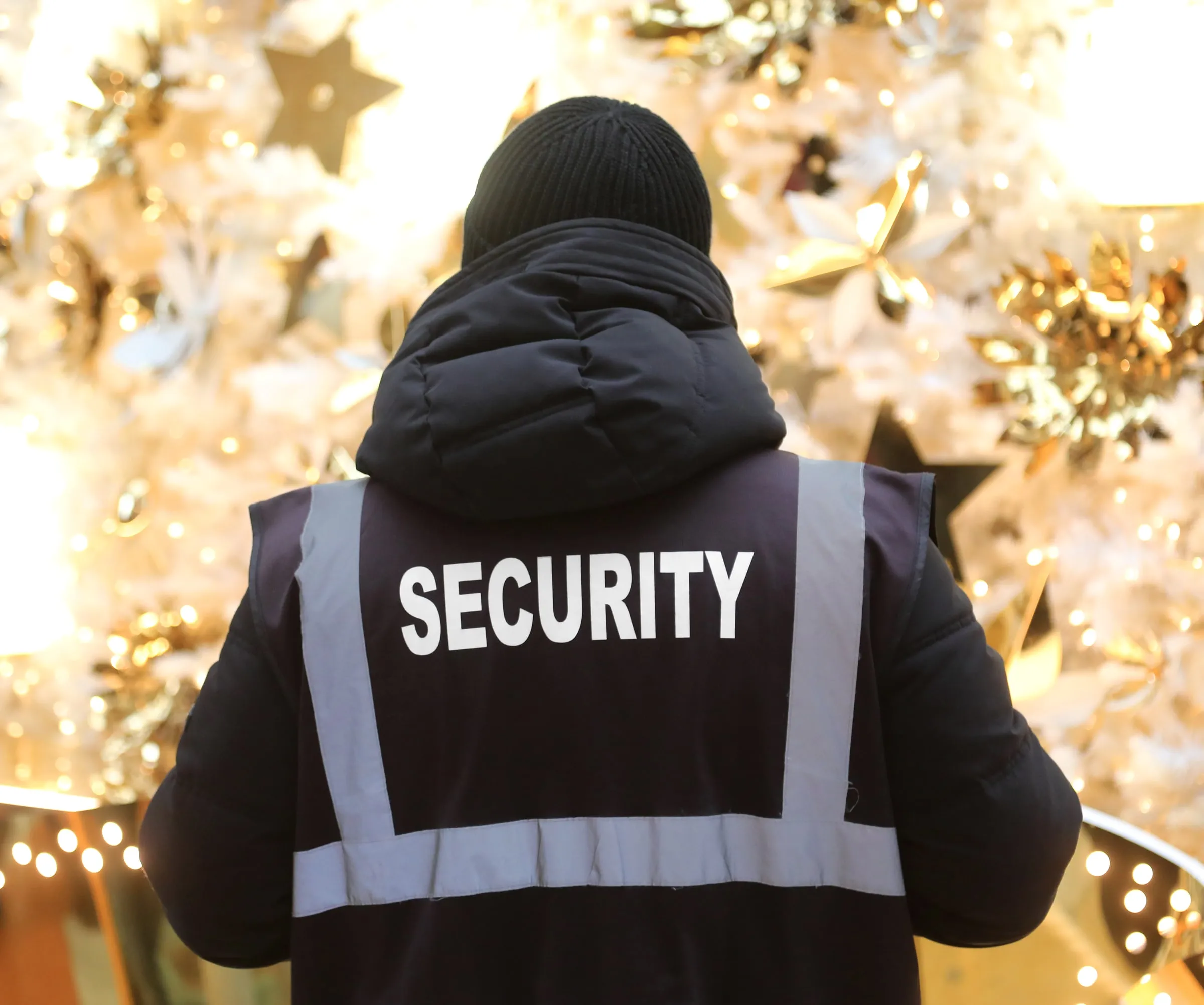Officer in a navy SECURITY vest and knit cap facing a lit white and gold holiday display