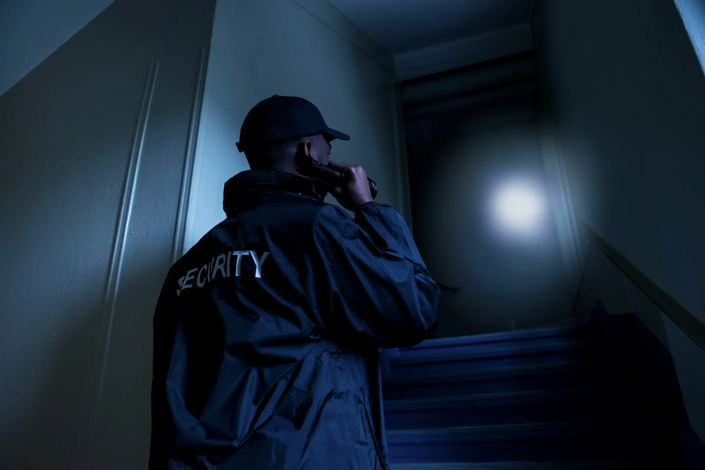 Low angle of an officer in a dark SECURITY jacket and cap aiming a flashlight up an unlit stairwell, the beam lighting the wall above