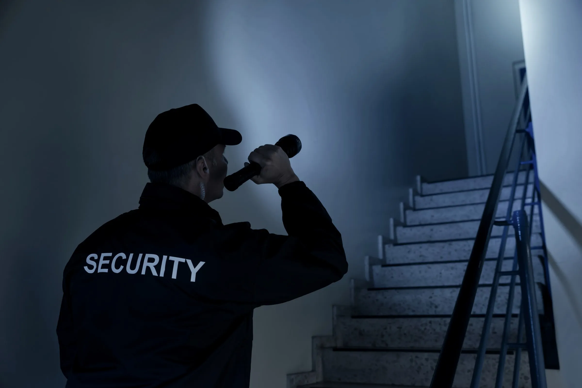 Officer seen from behind in a dark SECURITY jacket and cap shines a flashlight up a darkened stairwell beside a metal handrail