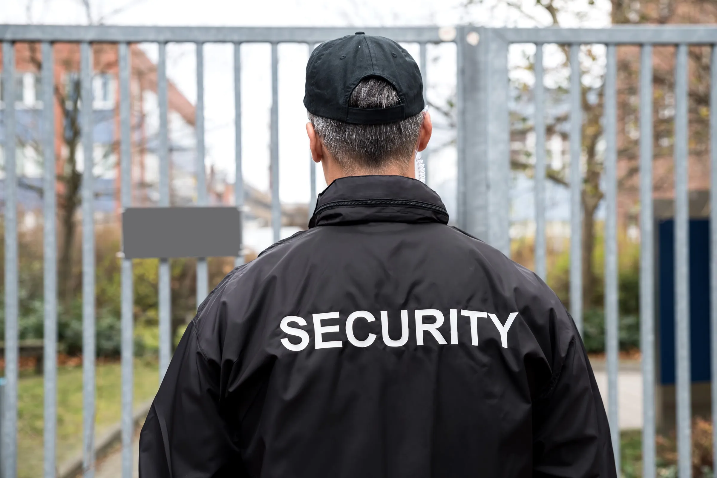 Security officer in a black SECURITY jacket and cap seen from behind at a white metal gate
