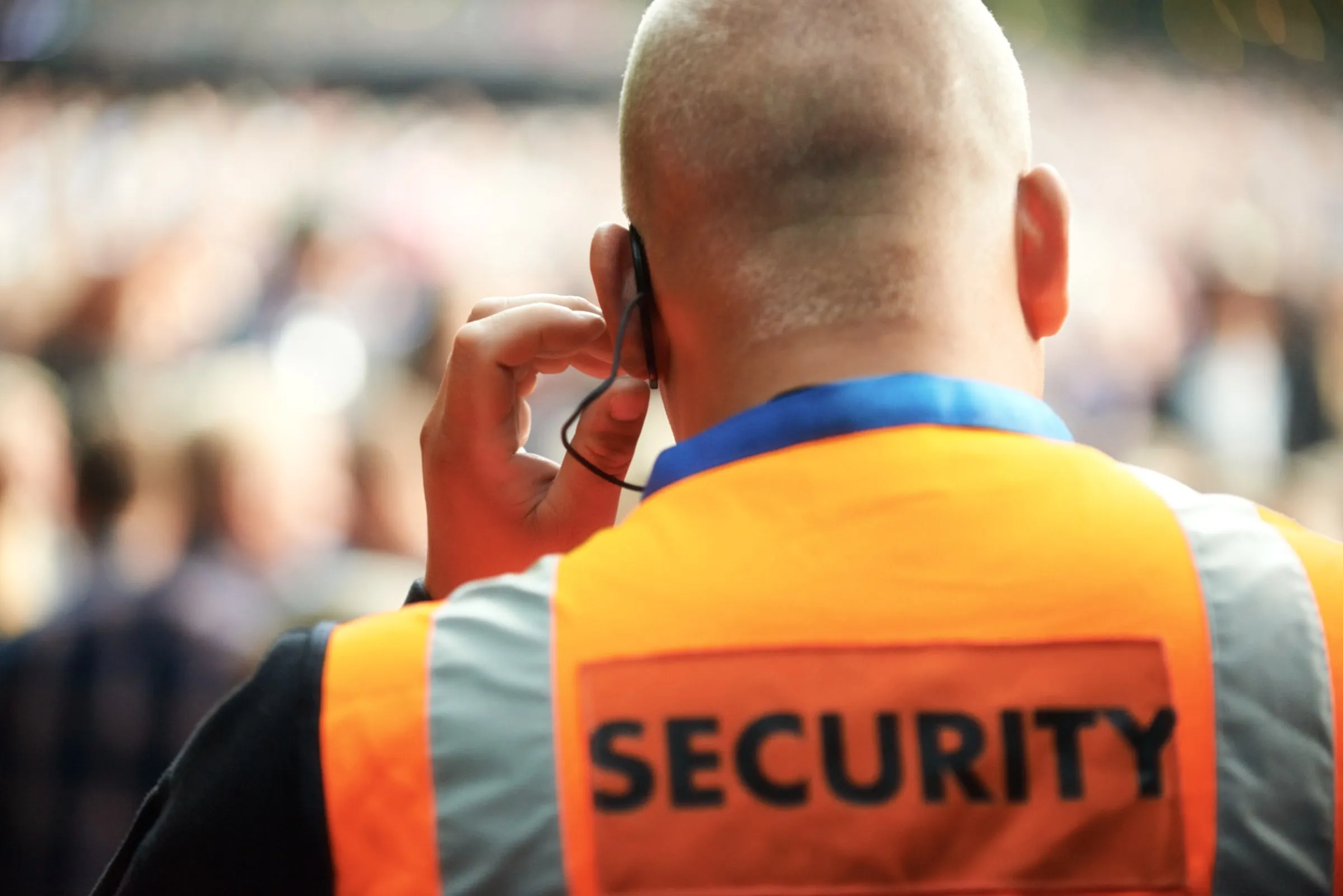 Officer in an orange high-visibility SECURITY vest adjusting an earpiece with his back to a blurred crowd