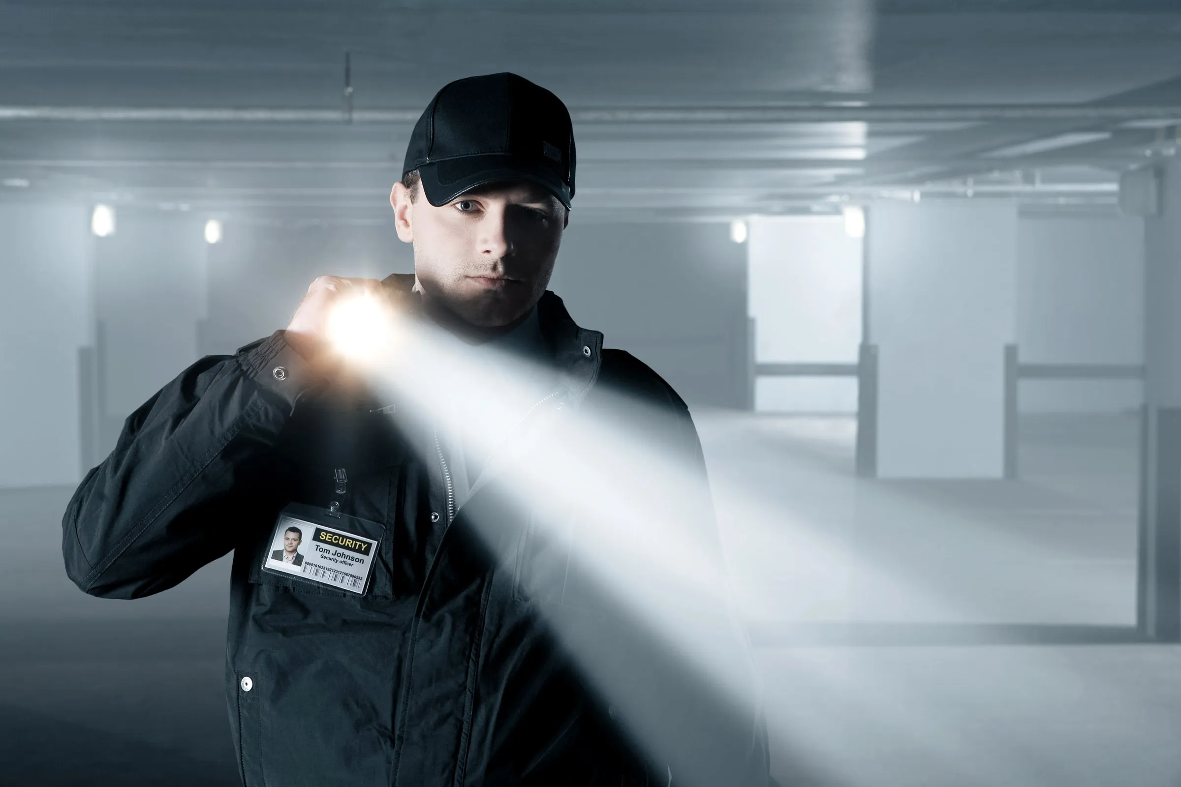 Security officer in a black cap and jacket with a SECURITY badge shining a flashlight across an empty parking garage