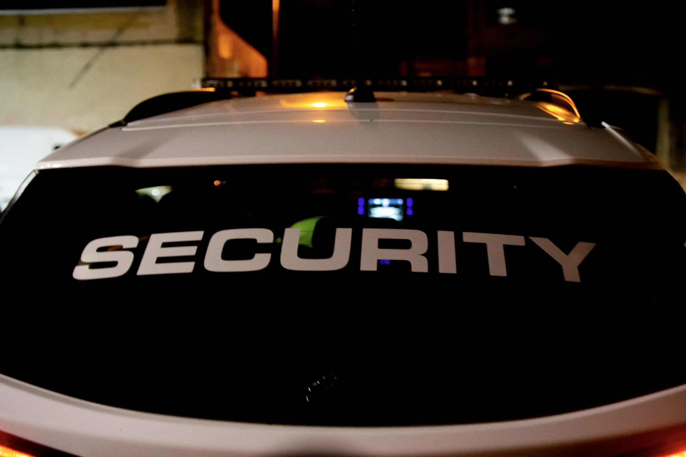 Rear window of a white patrol vehicle marked SECURITY with a roof light bar, parked at night