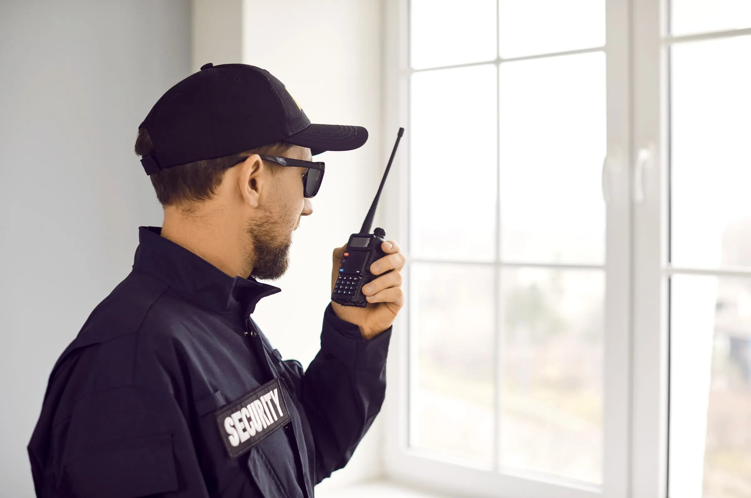 Side profile of a bearded officer in a dark uniform with a SECURITY patch, cap and sunglasses speaking into a handheld radio beside a bright window